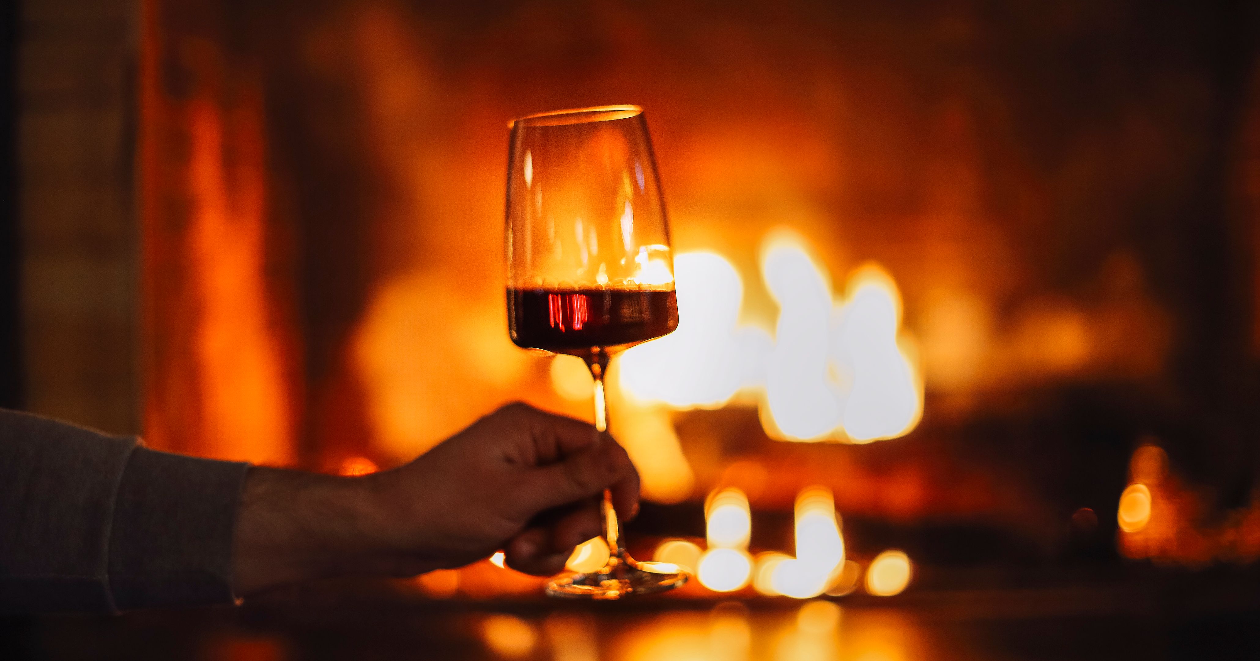 Hand holding a glass of red wine in front of a glowing fireplace