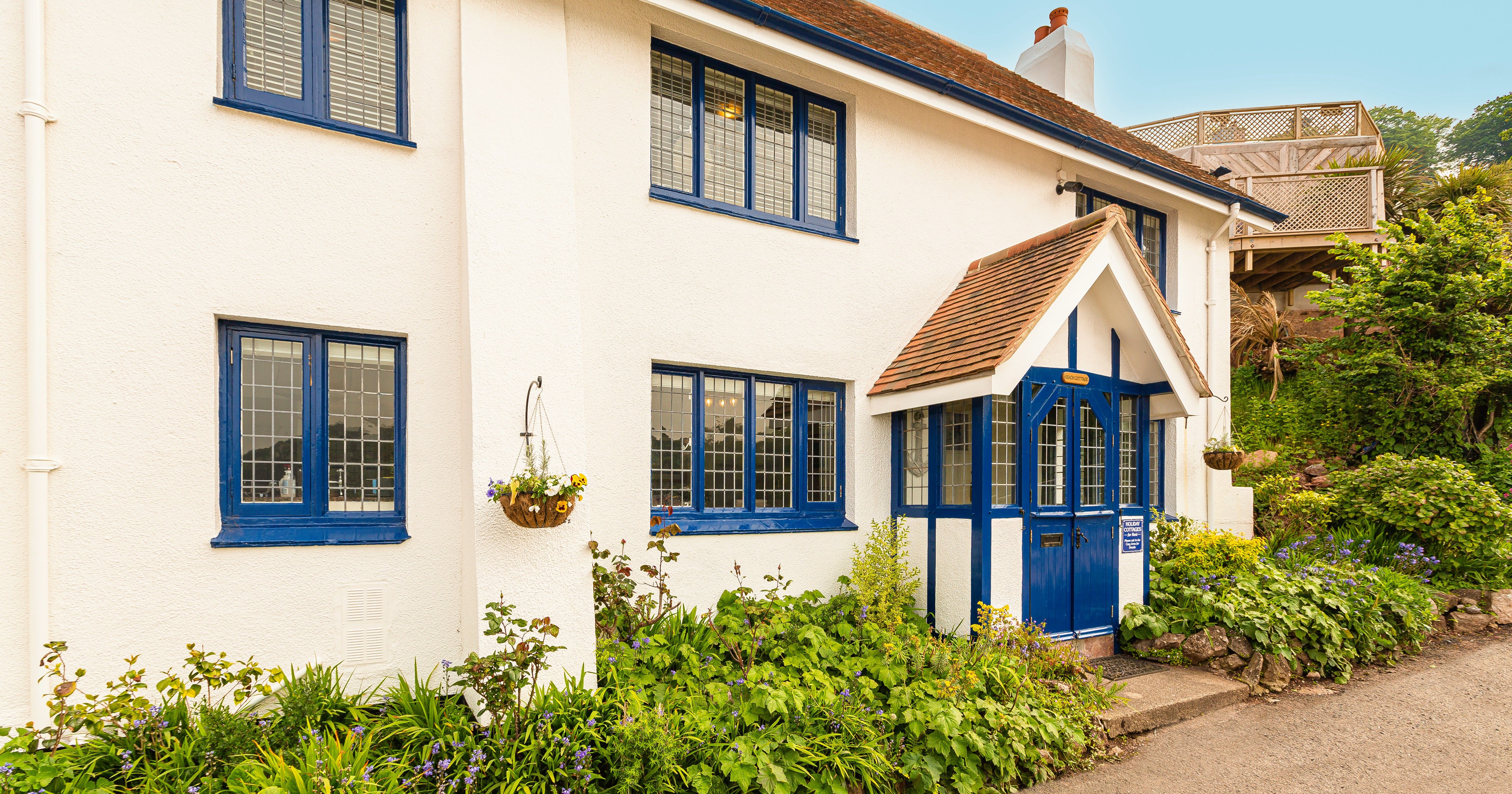 White cottage exterior with blue-framed windows and a blue front porch, surrounded by lush garden plants