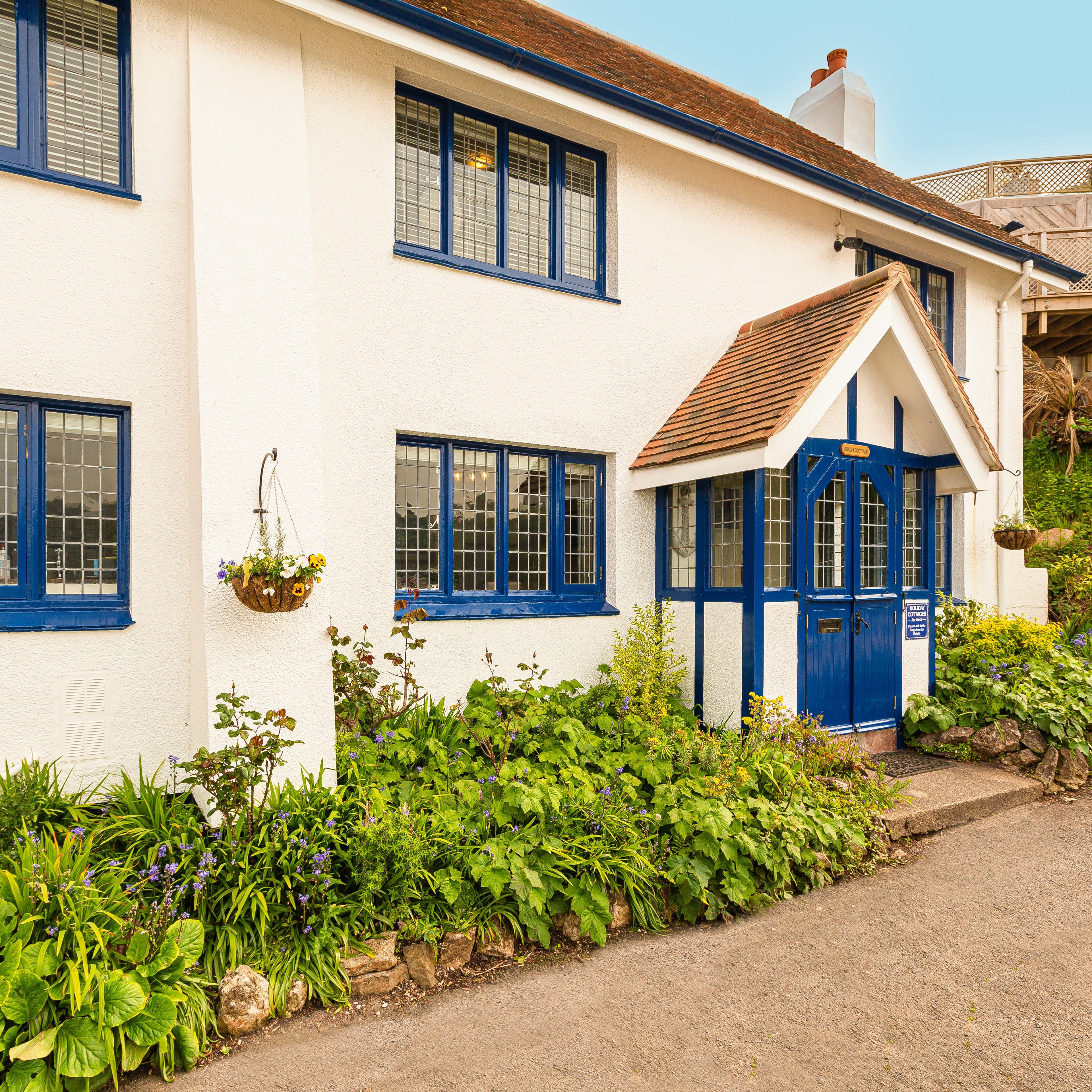White cottage exterior with blue-framed windows and a blue front porch, surrounded by lush garden plants