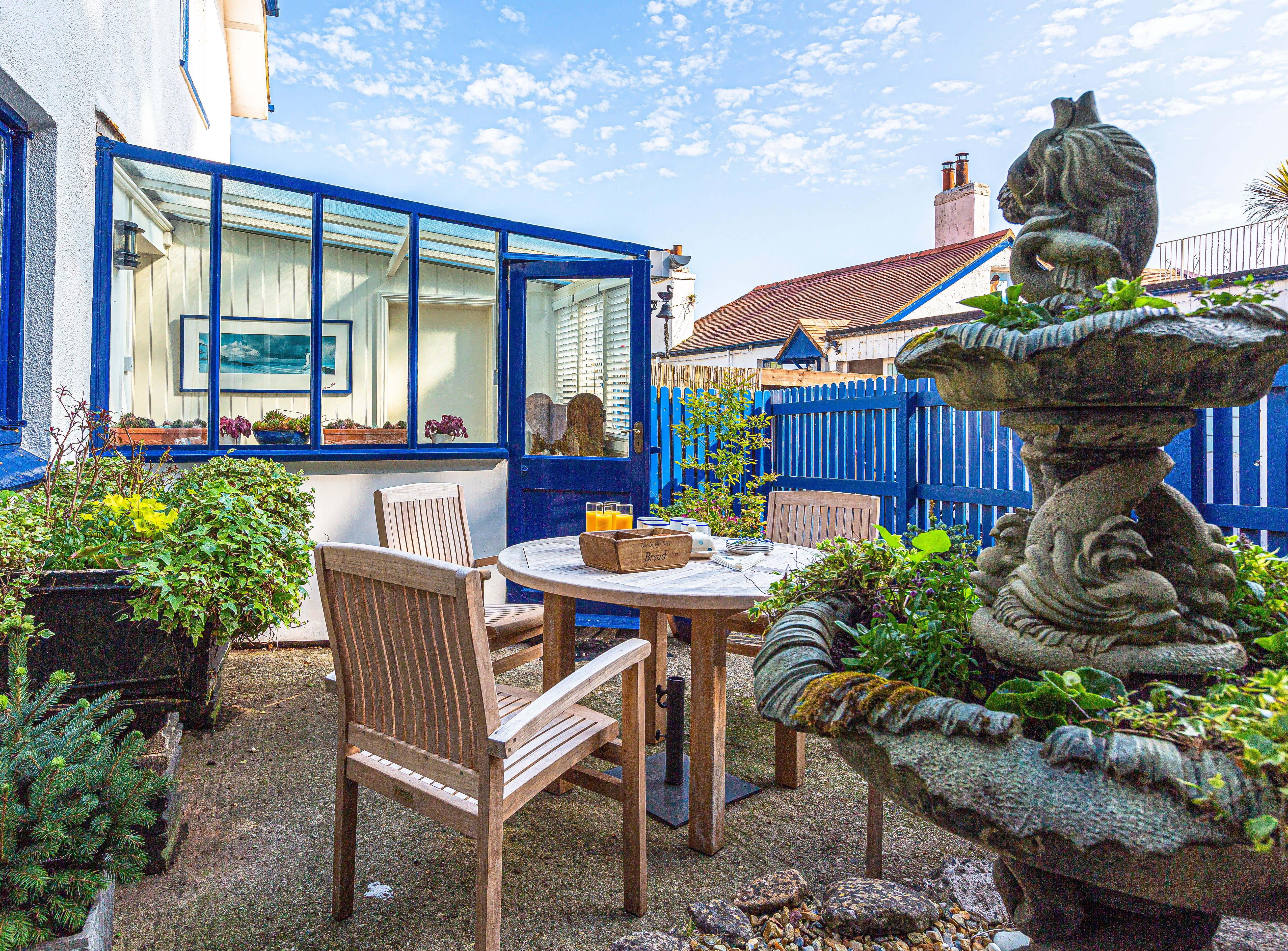 Sunlit patio with wooden table and chairs beside a blue-framed conservatory and garden fountain
