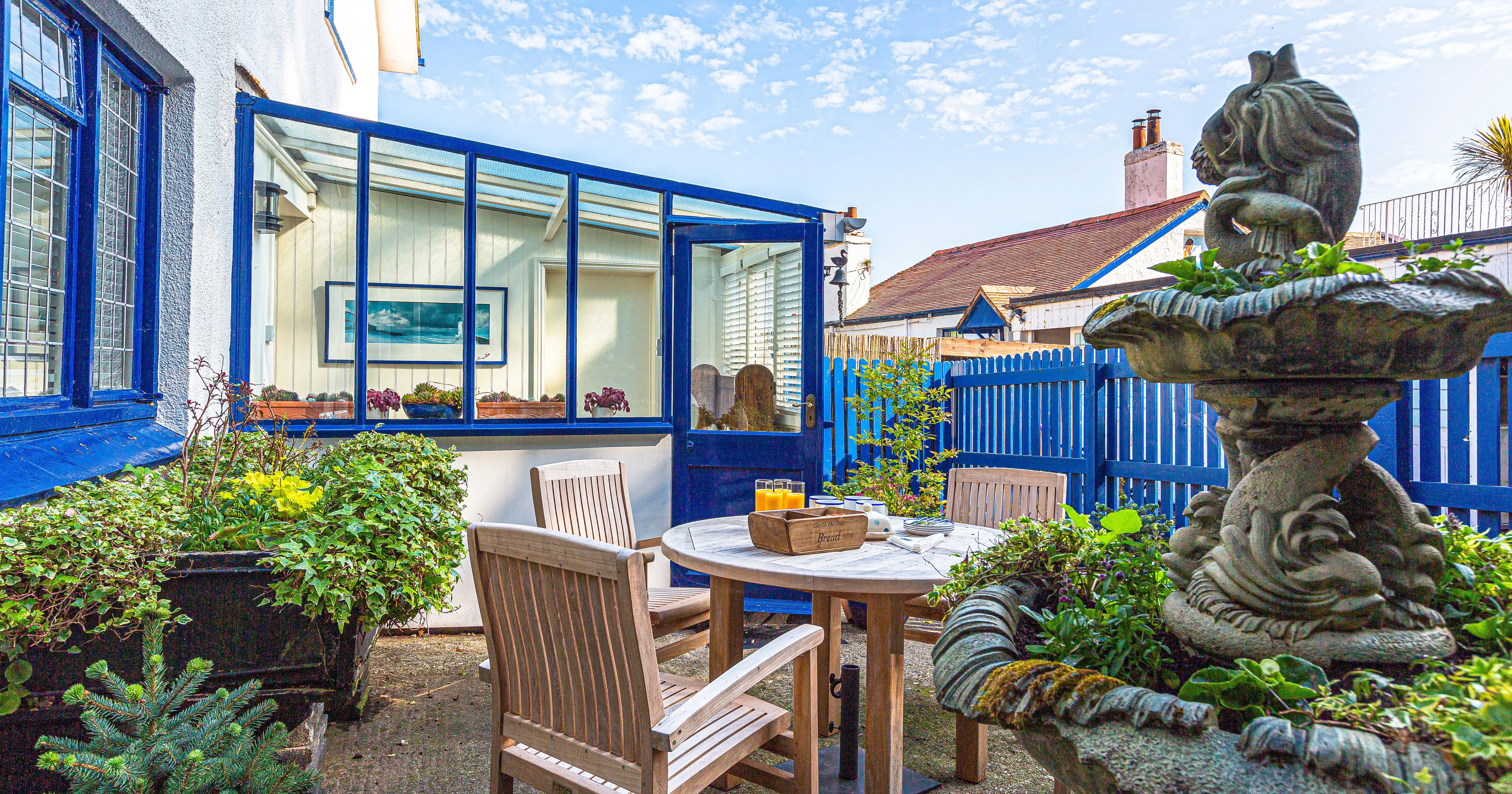 Sunlit patio with wooden table and chairs beside a blue-framed conservatory and garden fountain