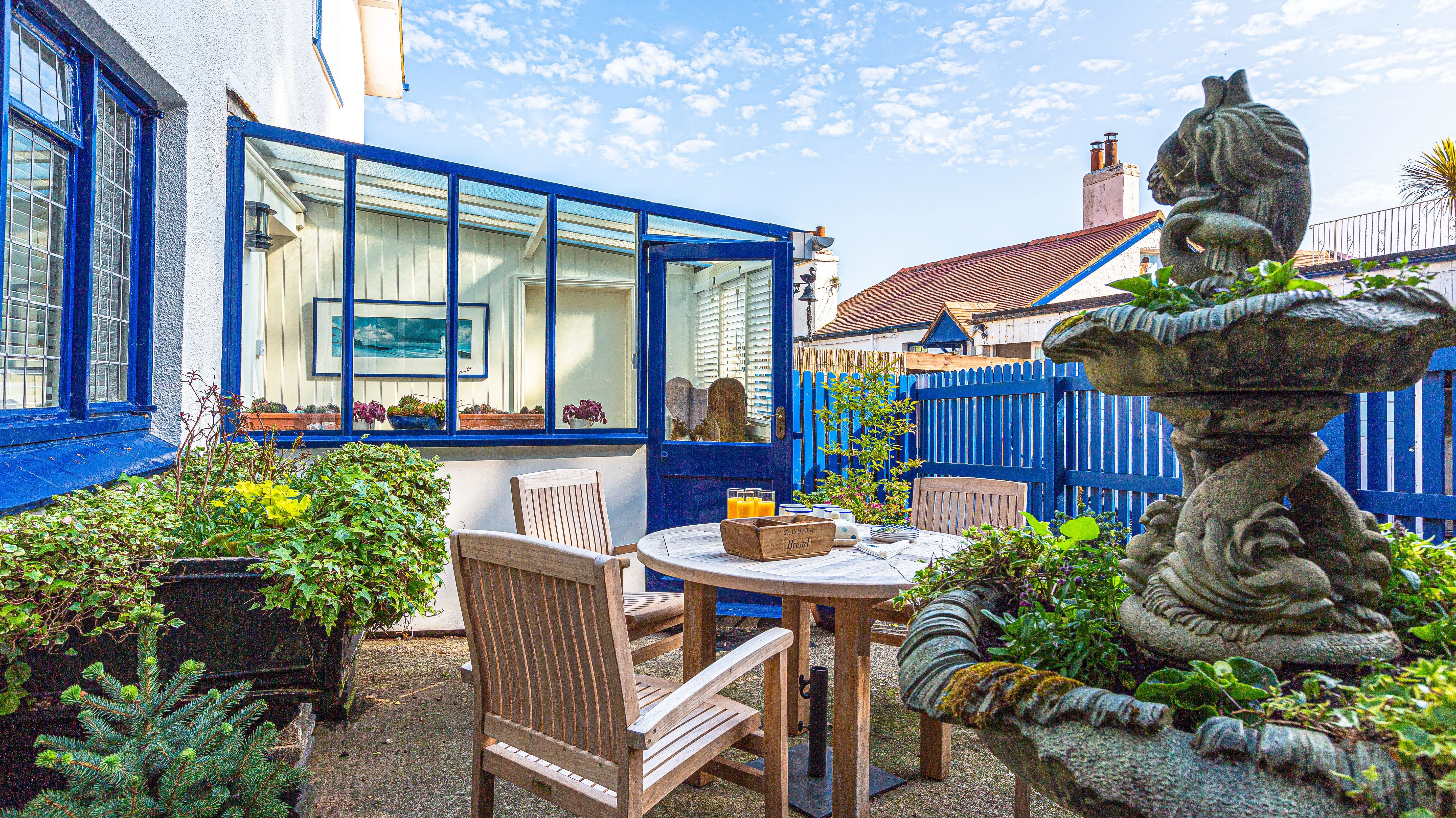 Sunlit patio with wooden table and chairs beside a blue-framed conservatory and garden fountain
