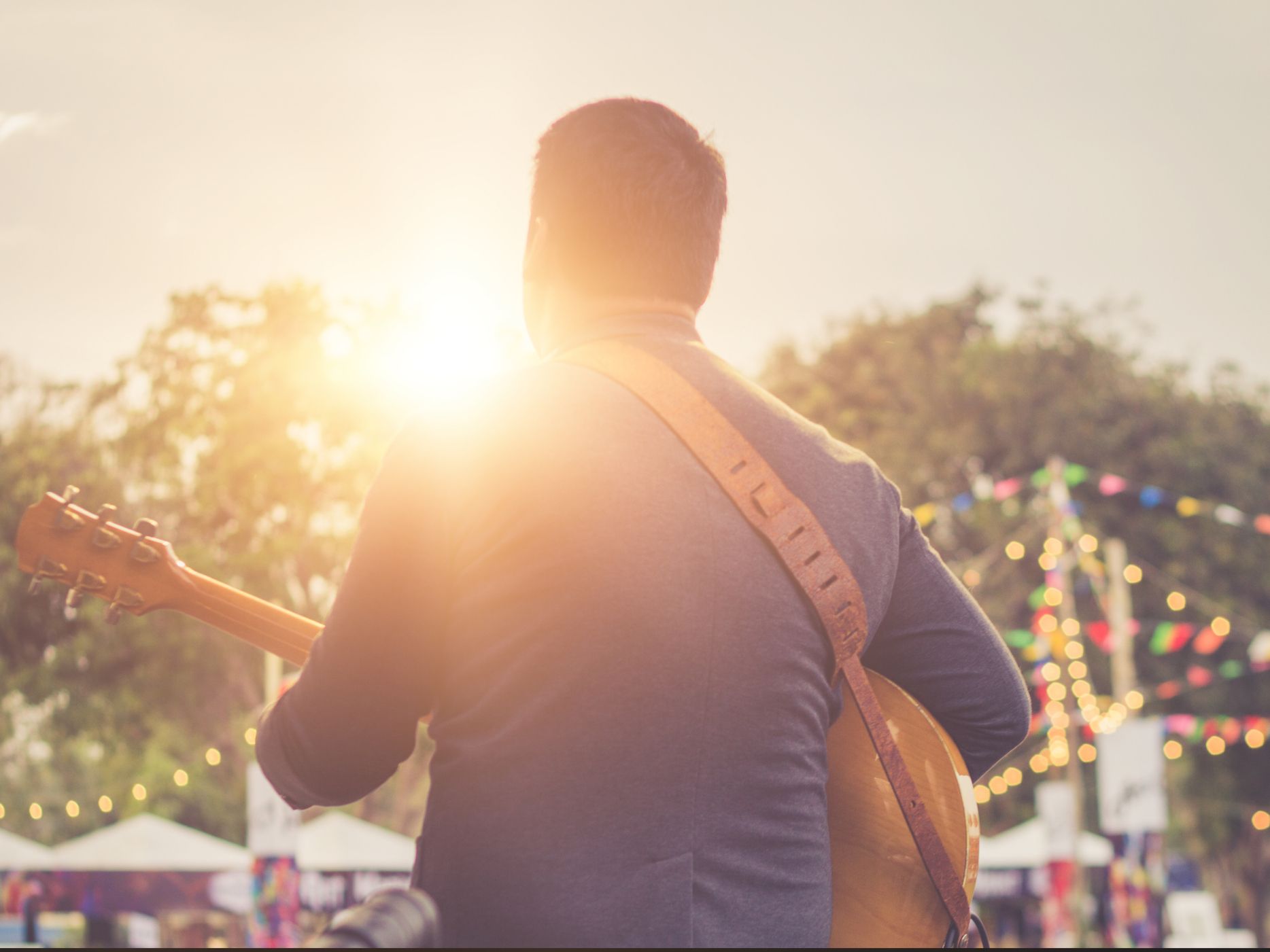Back view of a musician playing an acoustic guitar outdoors at sunset with festival lights and bunting