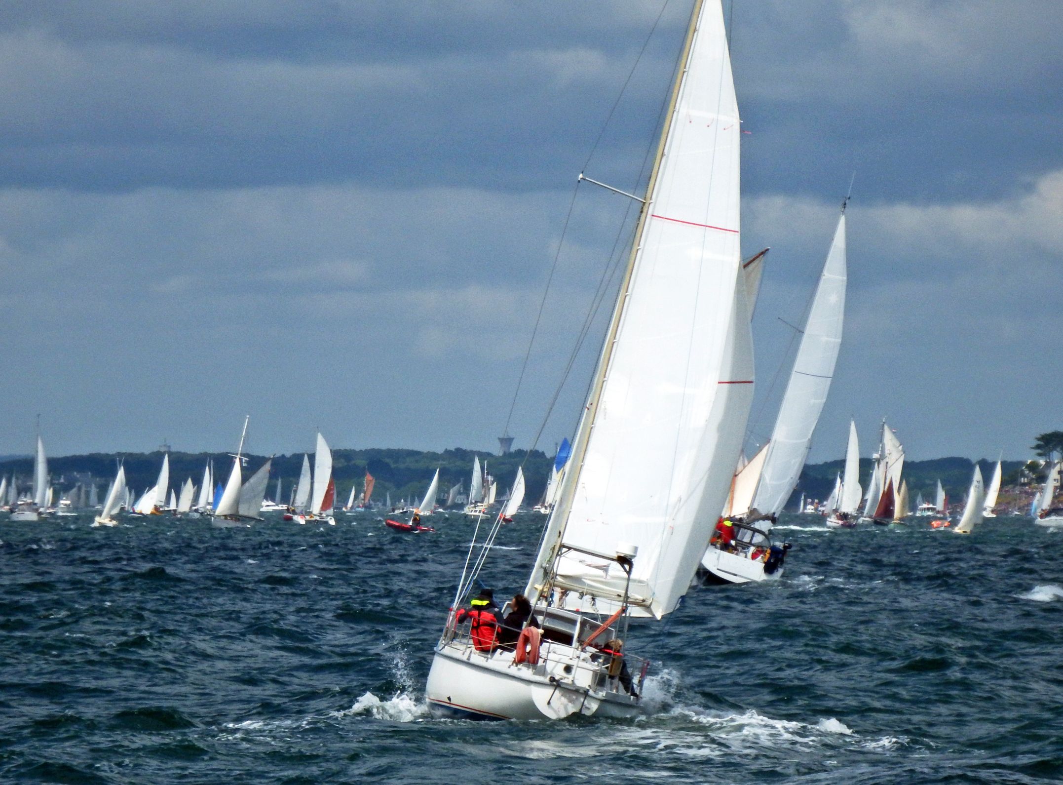 Sailing yacht heeling on choppy sea during a regatta, with many boats in the distance under a dark cloudy sky
