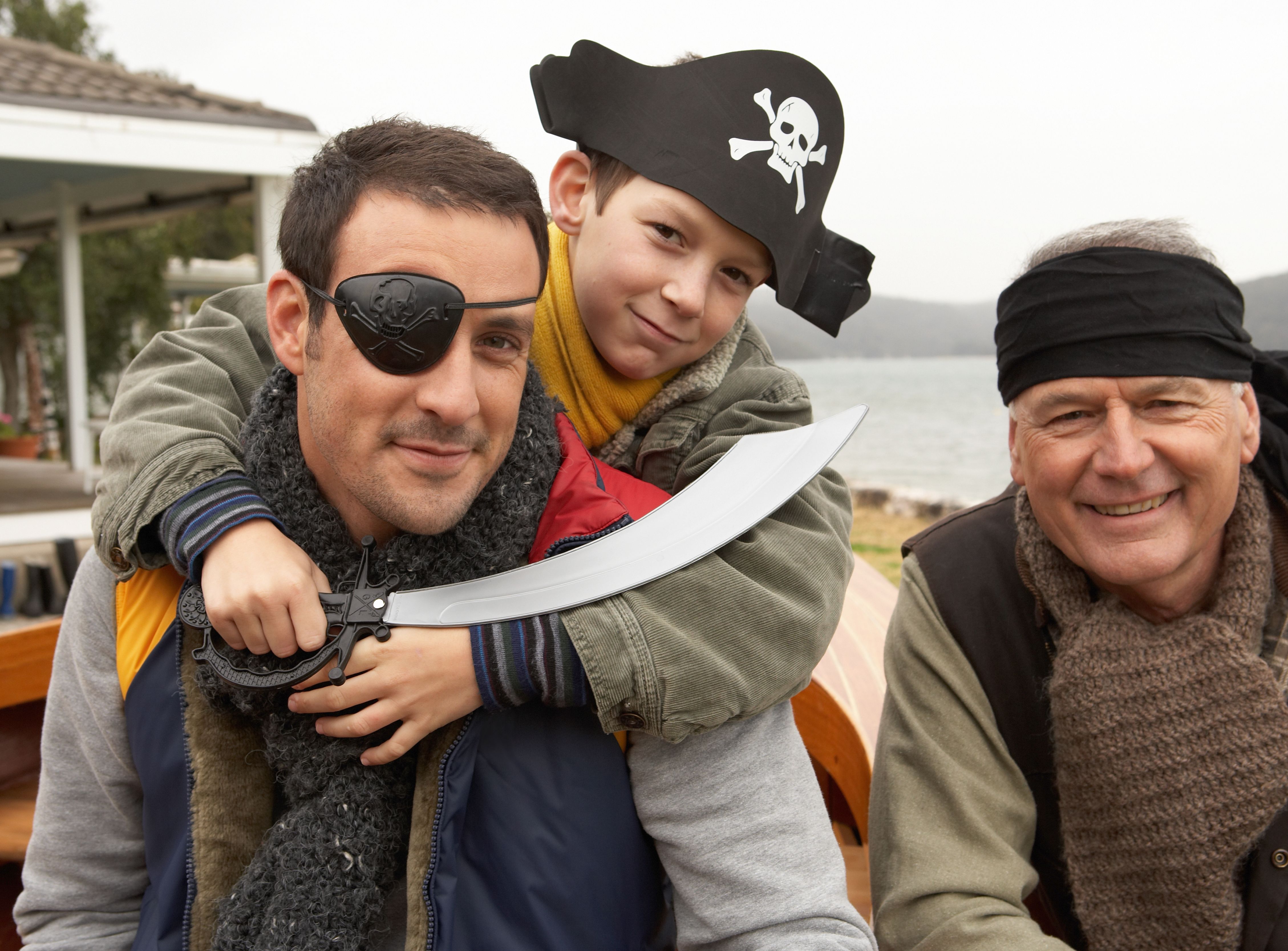 Boy dressed as a pirate hugs a man wearing an eye patch while an older man in a bandana smiles beside them outdoors.