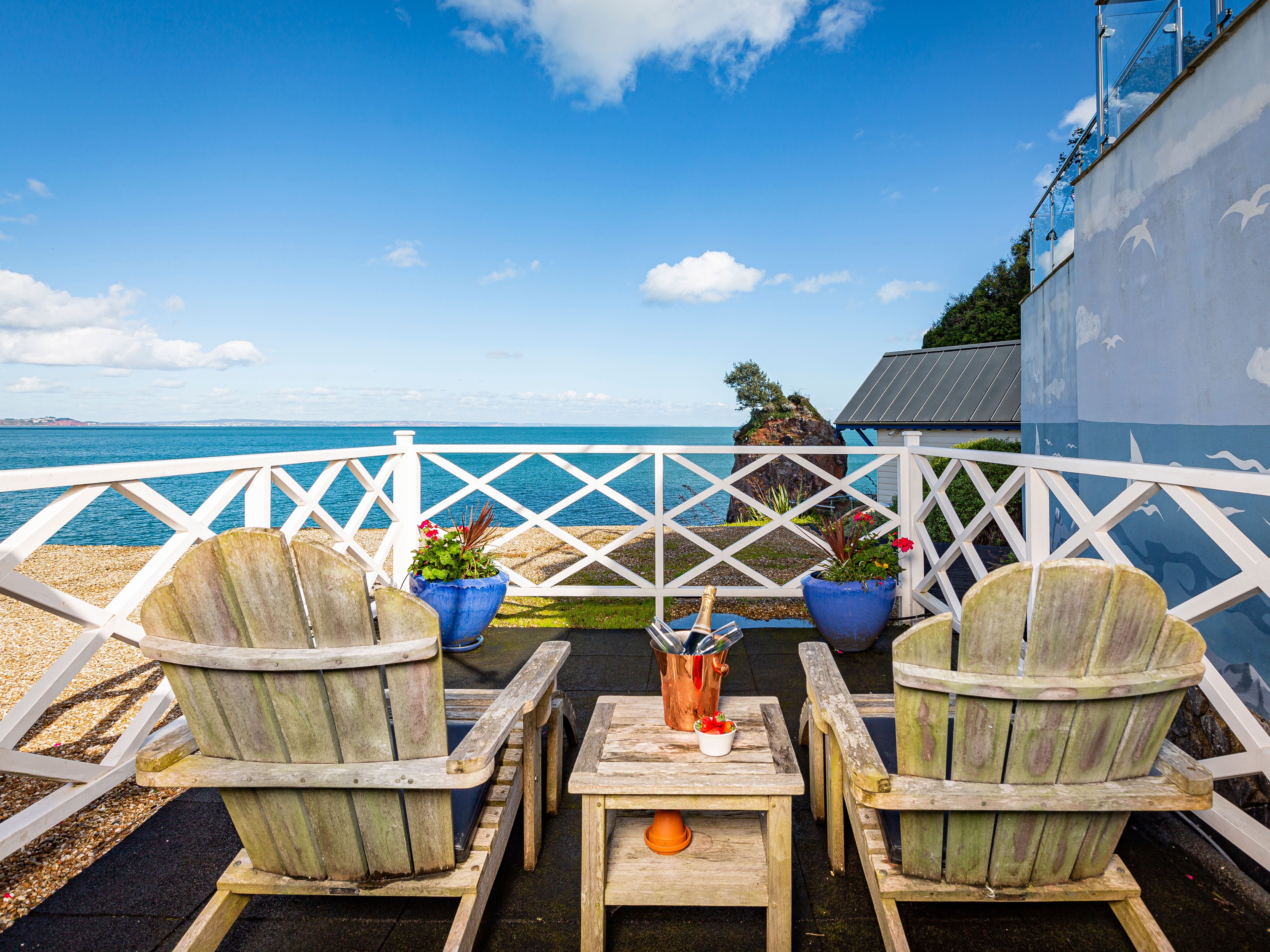 Seaside terrace with two wooden Adirondack chairs overlooking the sea, with a table holding an ice bucket and strawberries