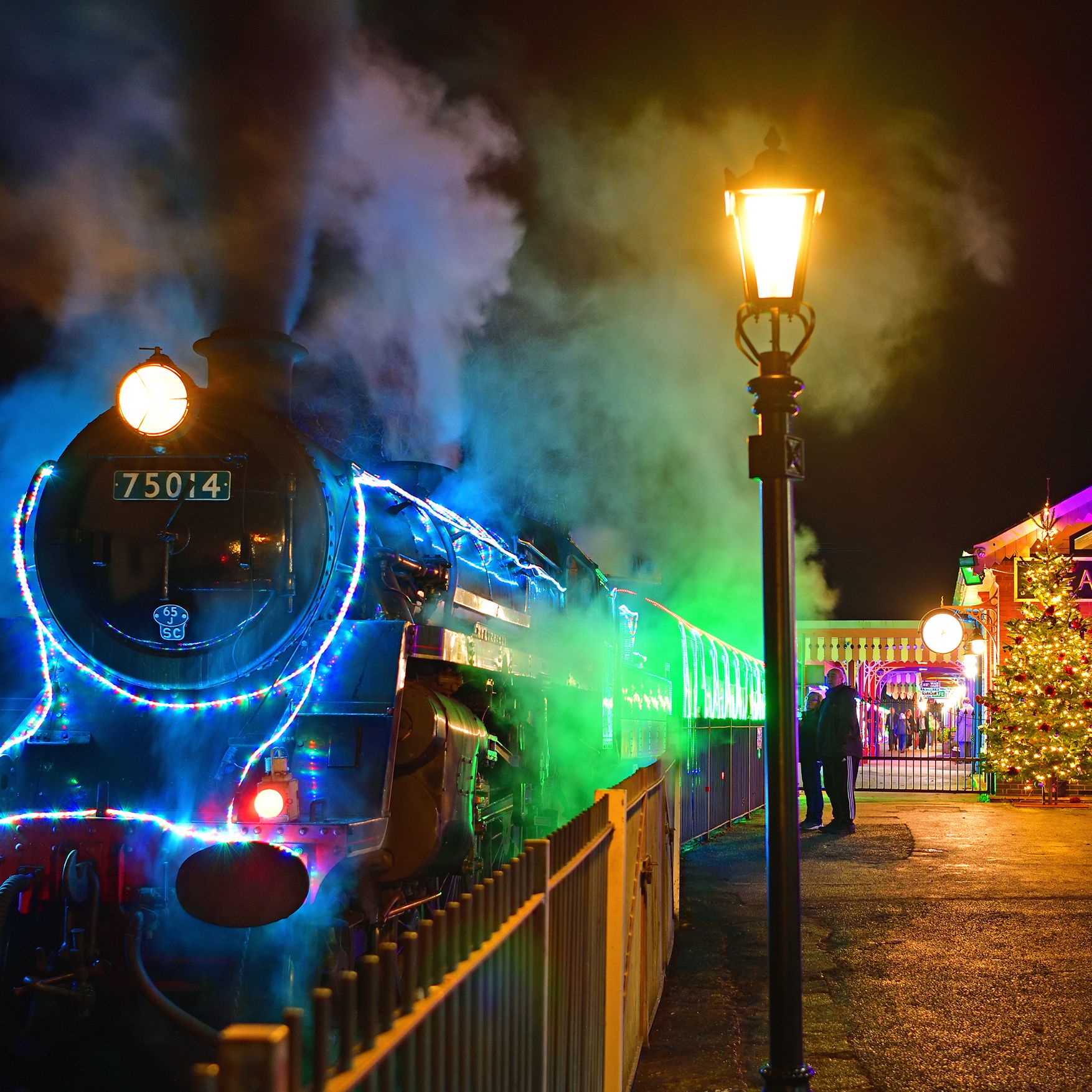 Steam locomotive decorated with colourful lights at a station platform at night, with steam rising beside the station entrance and a Christmas tree.