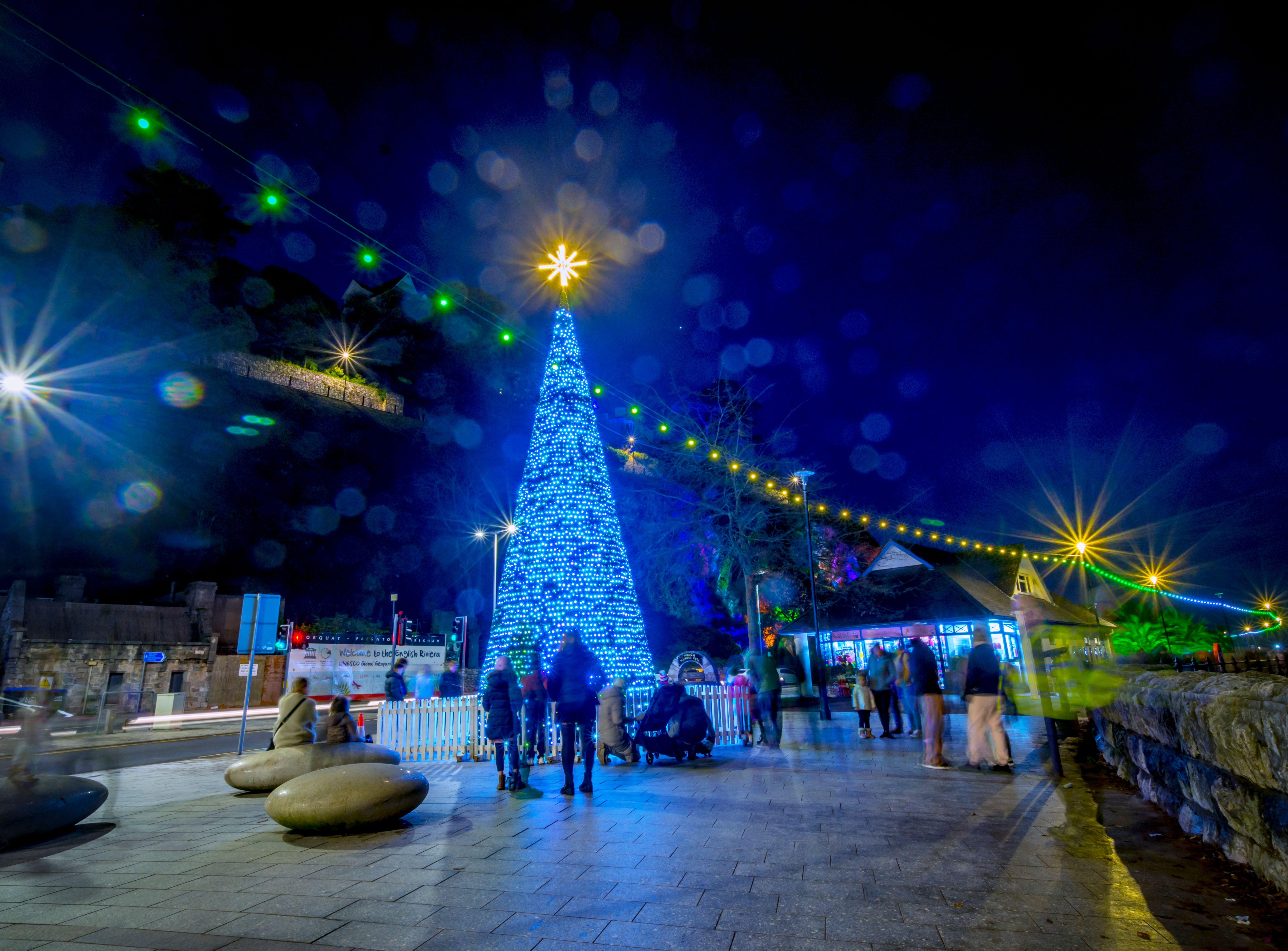 Blue illuminated Christmas tree with star topper at Bay of Lights festival at night, with festive string lights and people on the promenade