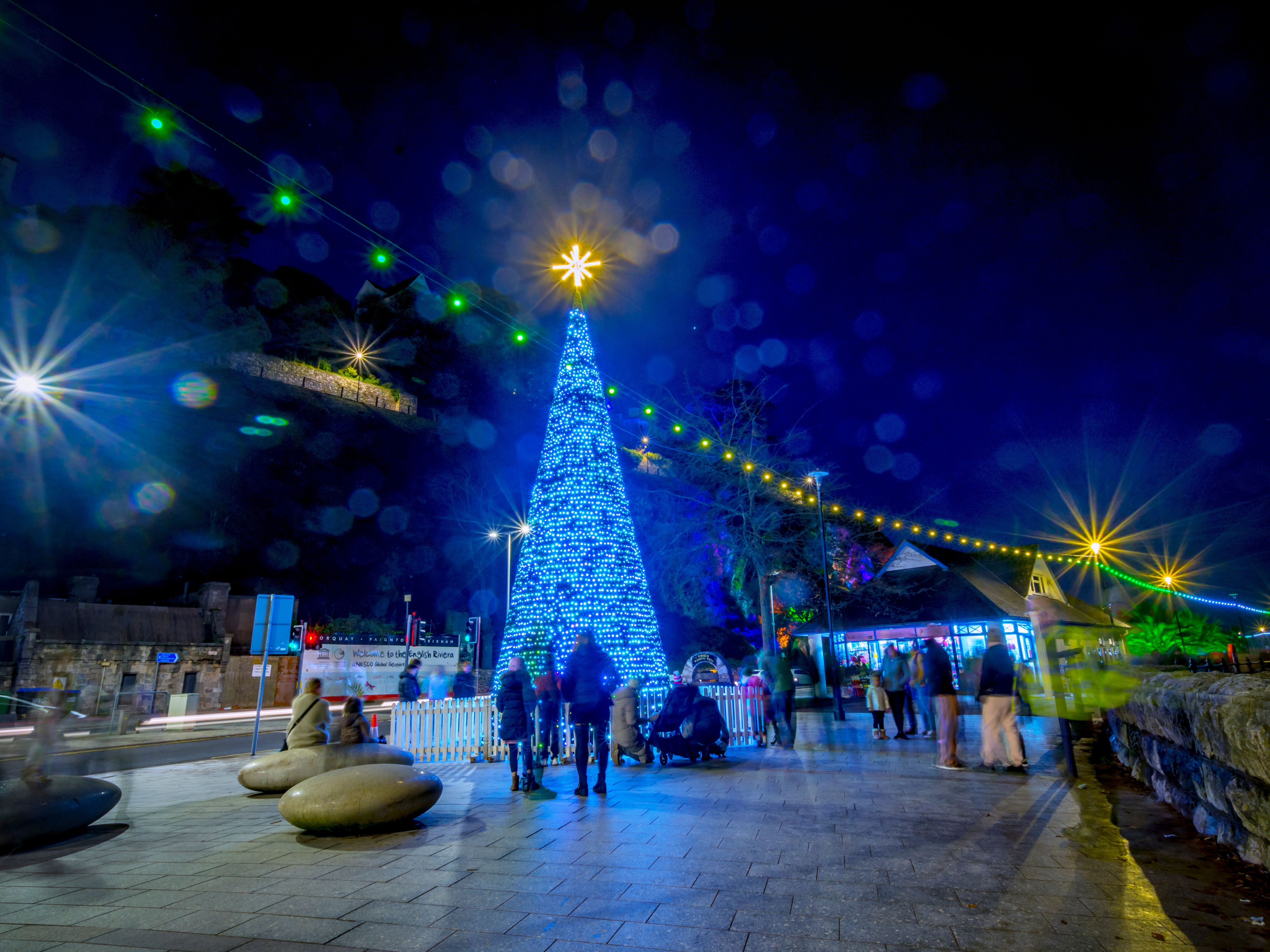Blue illuminated Christmas tree with star topper at Bay of Lights festival at night, with festive string lights and people on the promenade