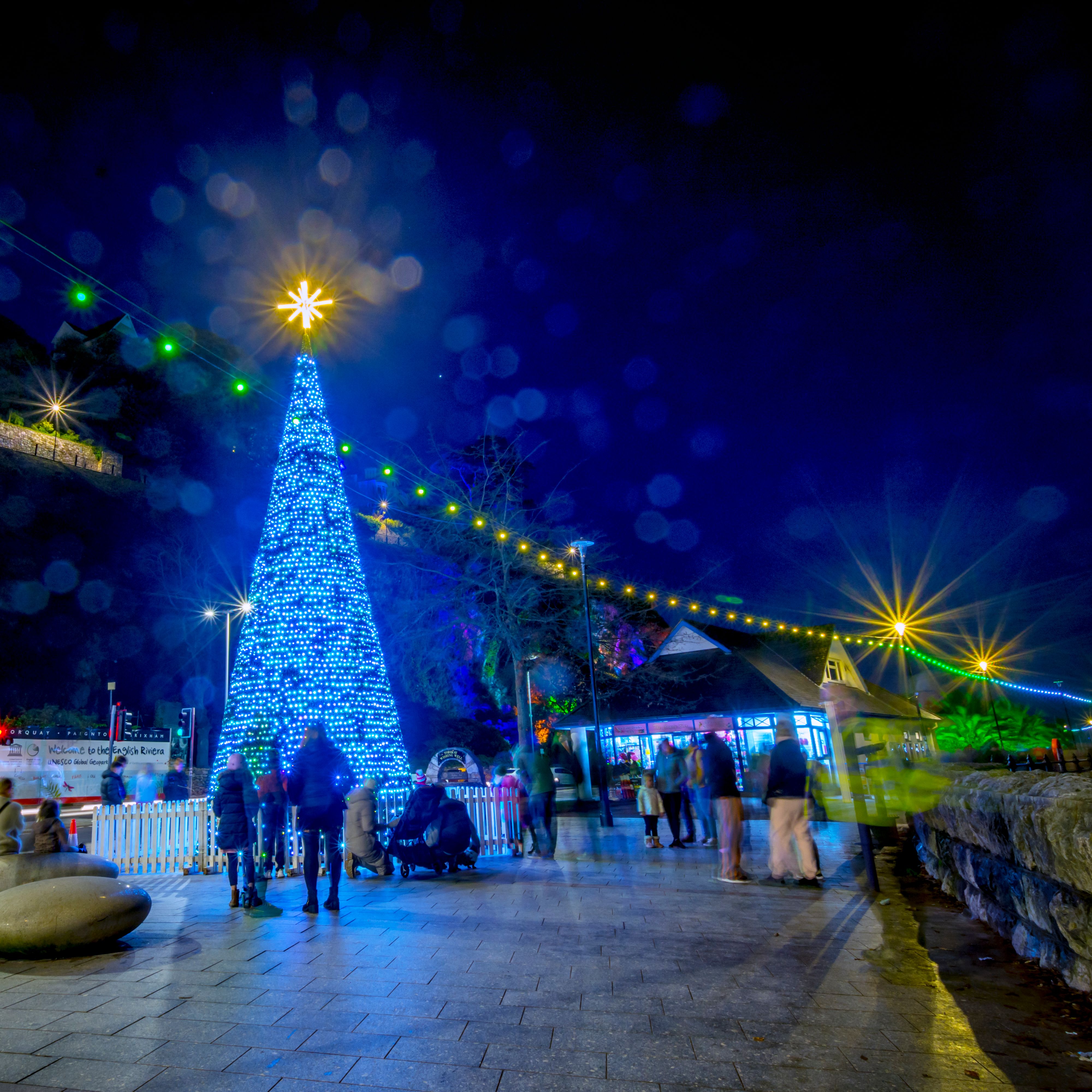 Blue illuminated Christmas tree with star topper at Bay of Lights festival at night, with festive string lights and people on the promenade