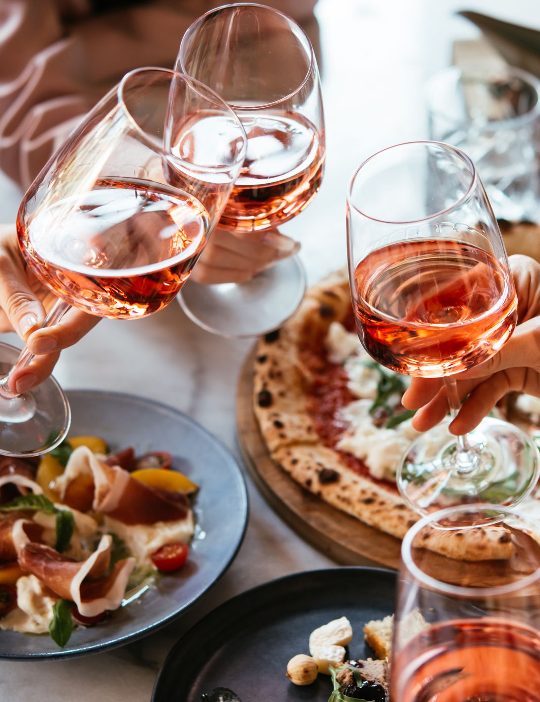 People toasting with glasses of rosé wine over a table with pizza and appetizers.