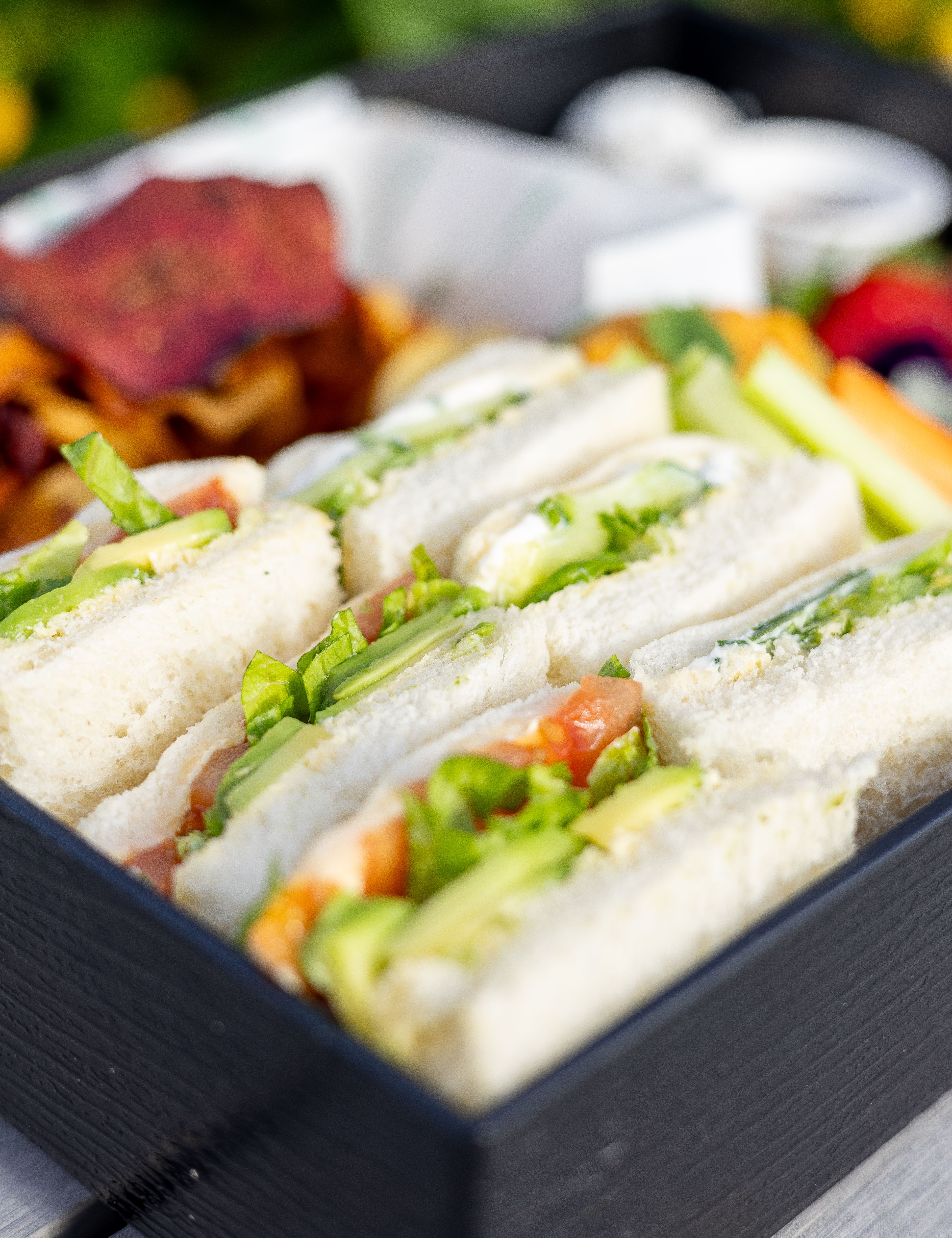 Vegetable tea sandwiches served in a black bento box with assorted chips and fresh vegetables.