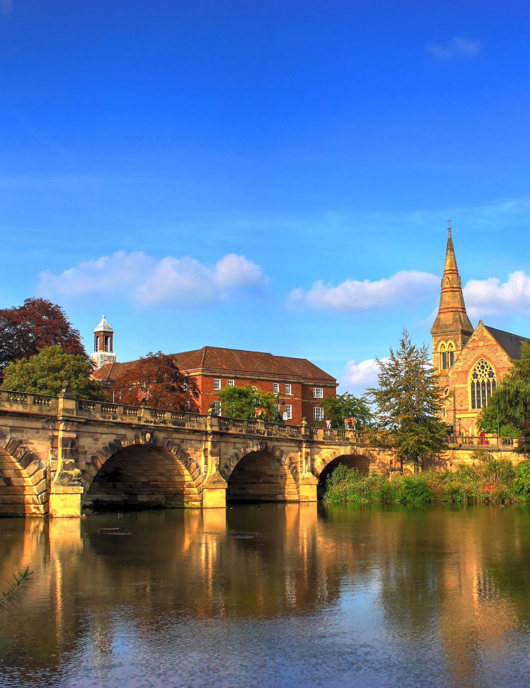 Historic stone bridge and church beside a river in a lush, green landscape under a blue sky.