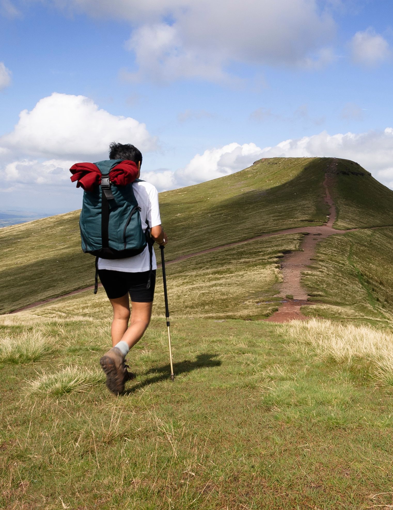 Hiker with backpack walking along a grassy mountain trail under a blue sky.