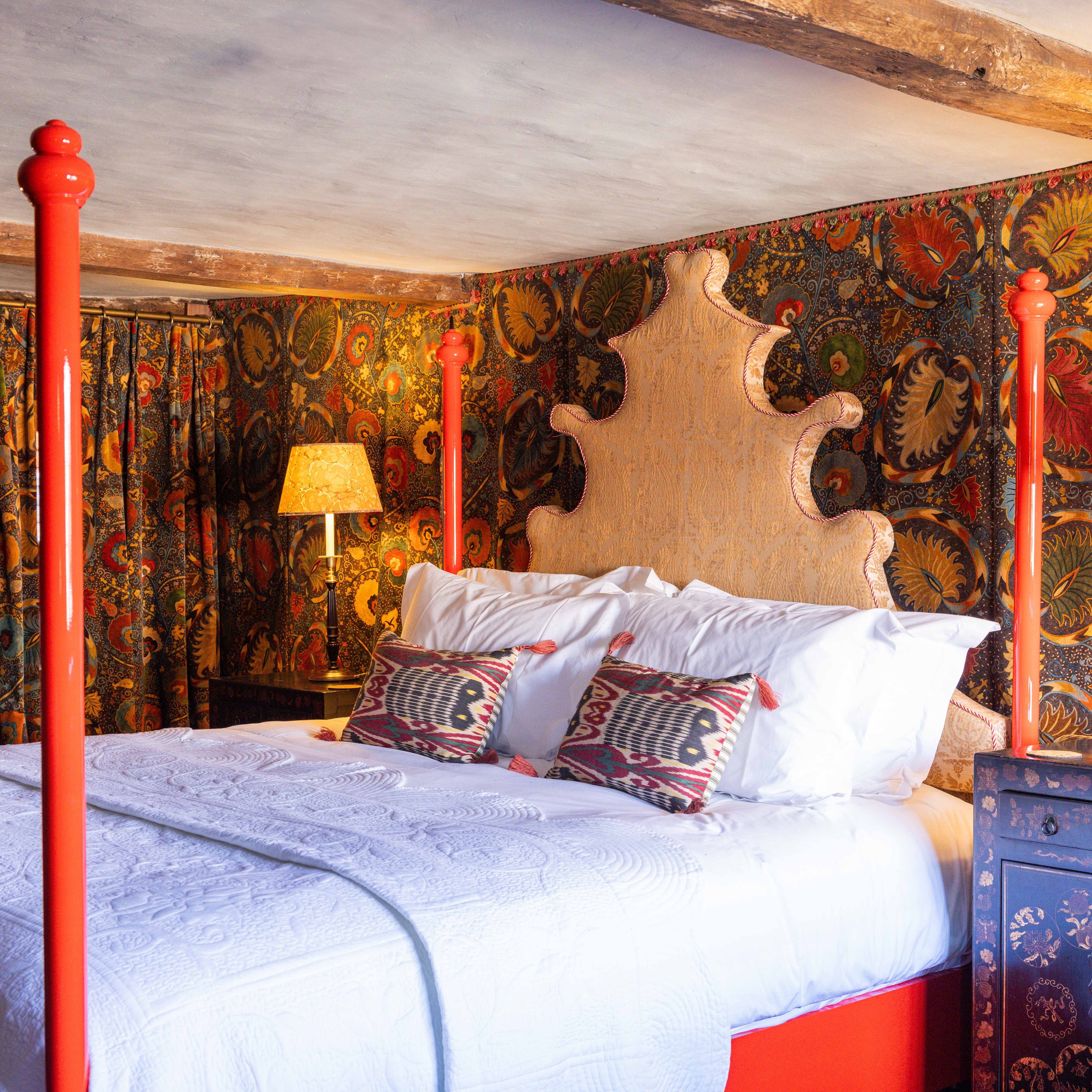 Ornate bed with red posts in a vintage-themed bedroom with patterned wallpaper and lamps.