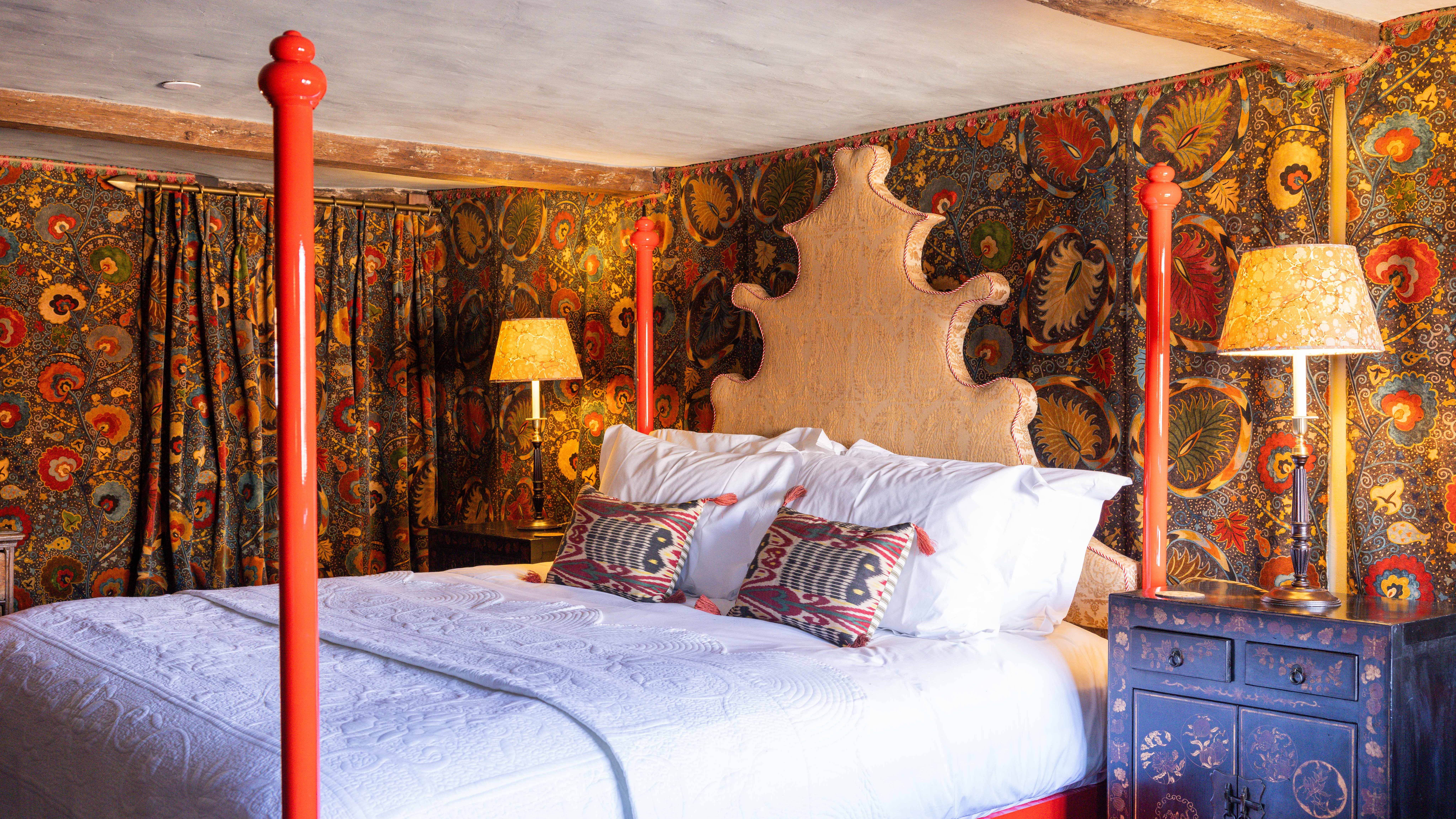 Ornate bed with red posts in a vintage-themed bedroom with patterned wallpaper and lamps.