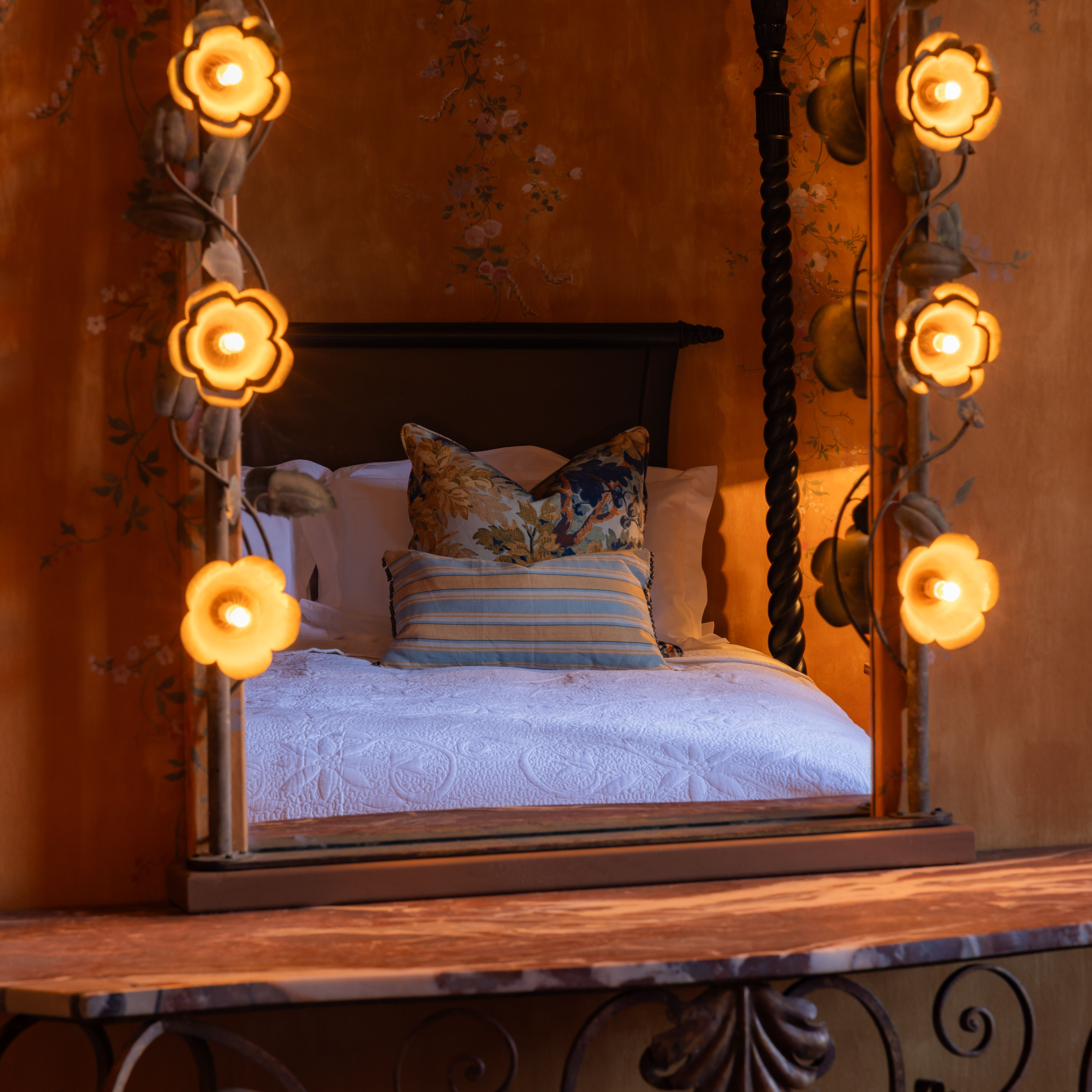 Ornate mirror with flower-shaped lights reflecting a cozy bed with decorative pillows and floral wallpaper