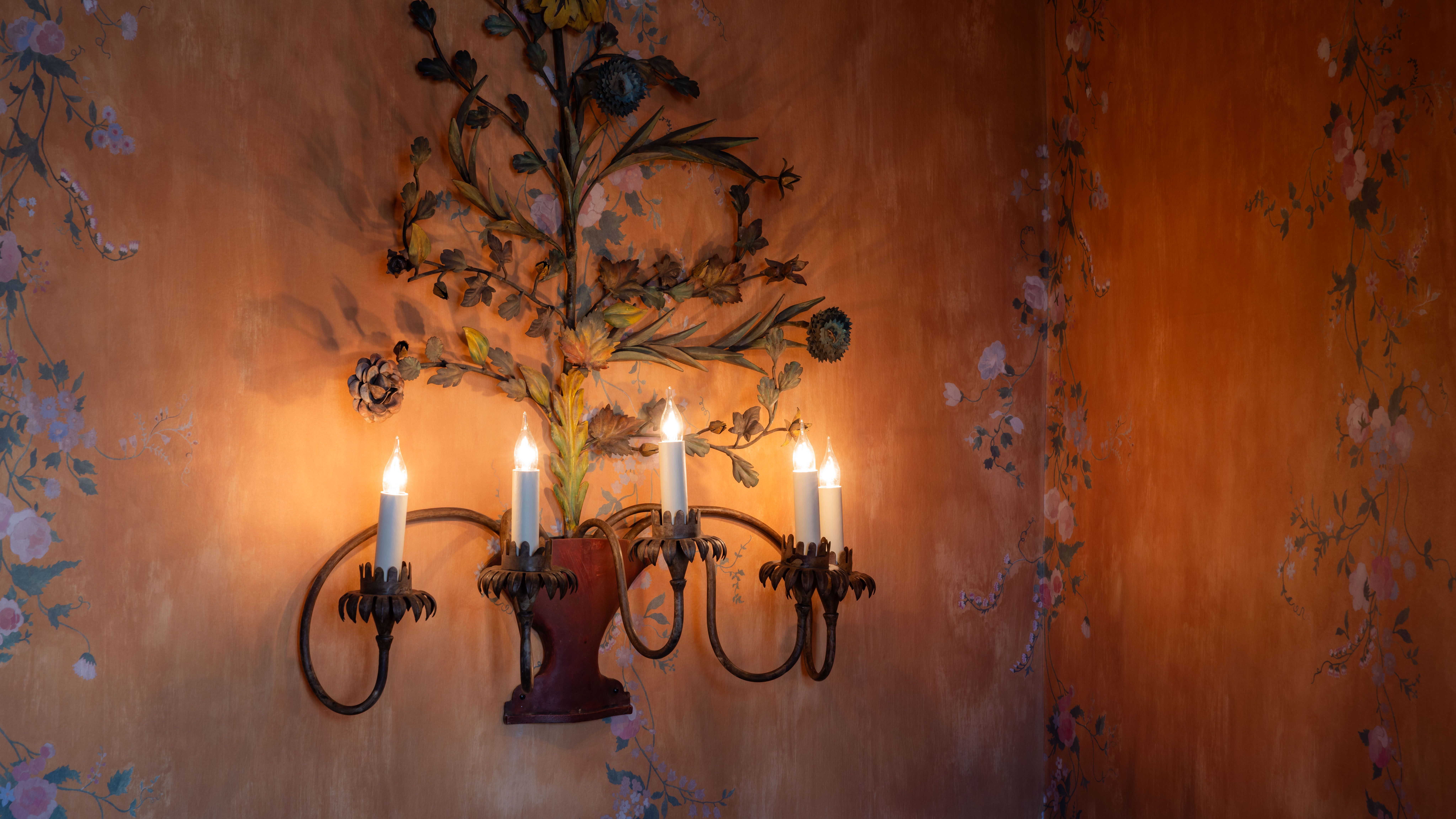 Decorative wall light with four candle-style bulbs and floral metal design mounted on an orange wall with painted floral patterns.