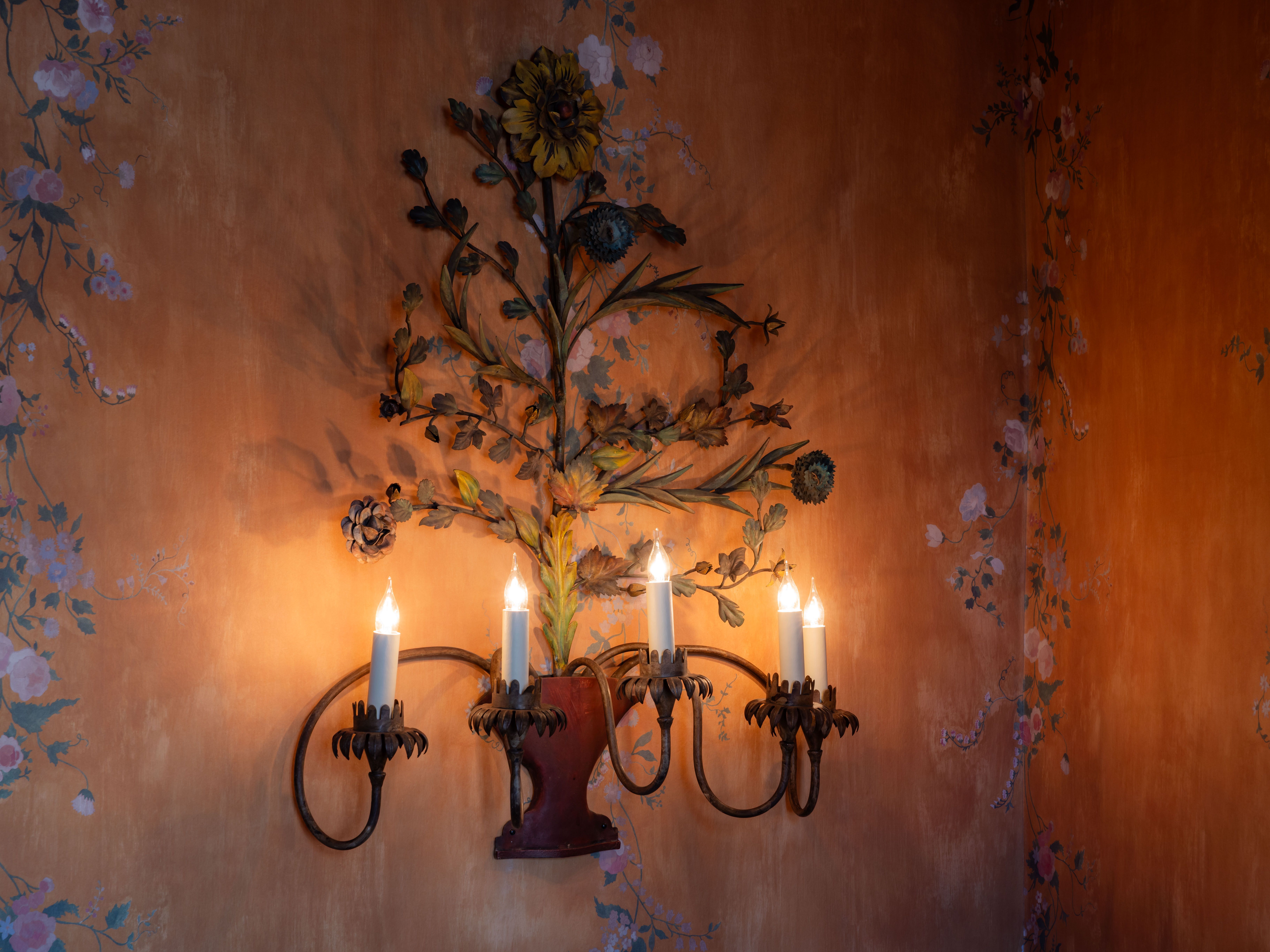Decorative wall light with four candle-style bulbs and floral metal design mounted on an orange wall with painted floral patterns.