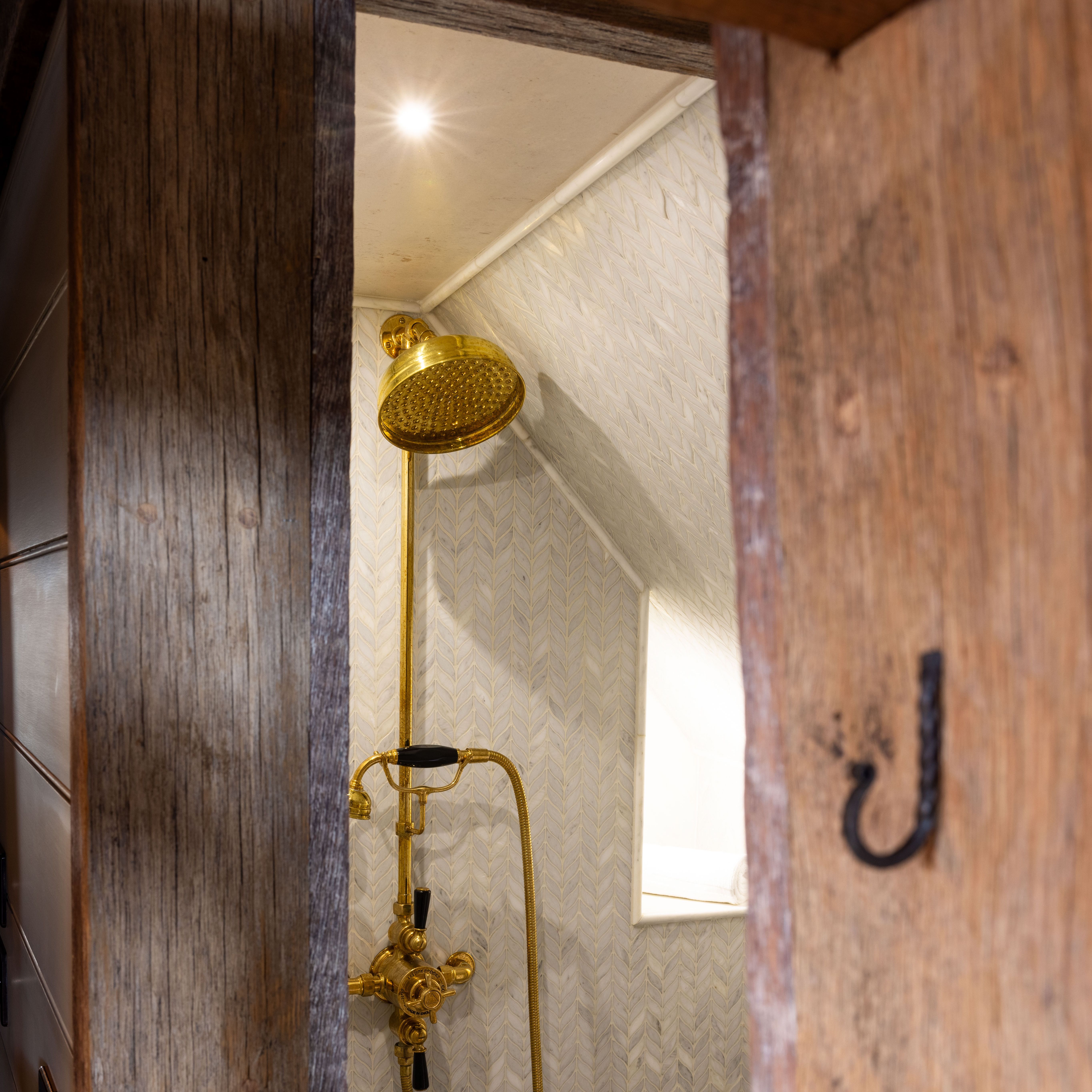 Vintage-style golden showerhead and controls inside a small tiled shower space with wooden door frame.