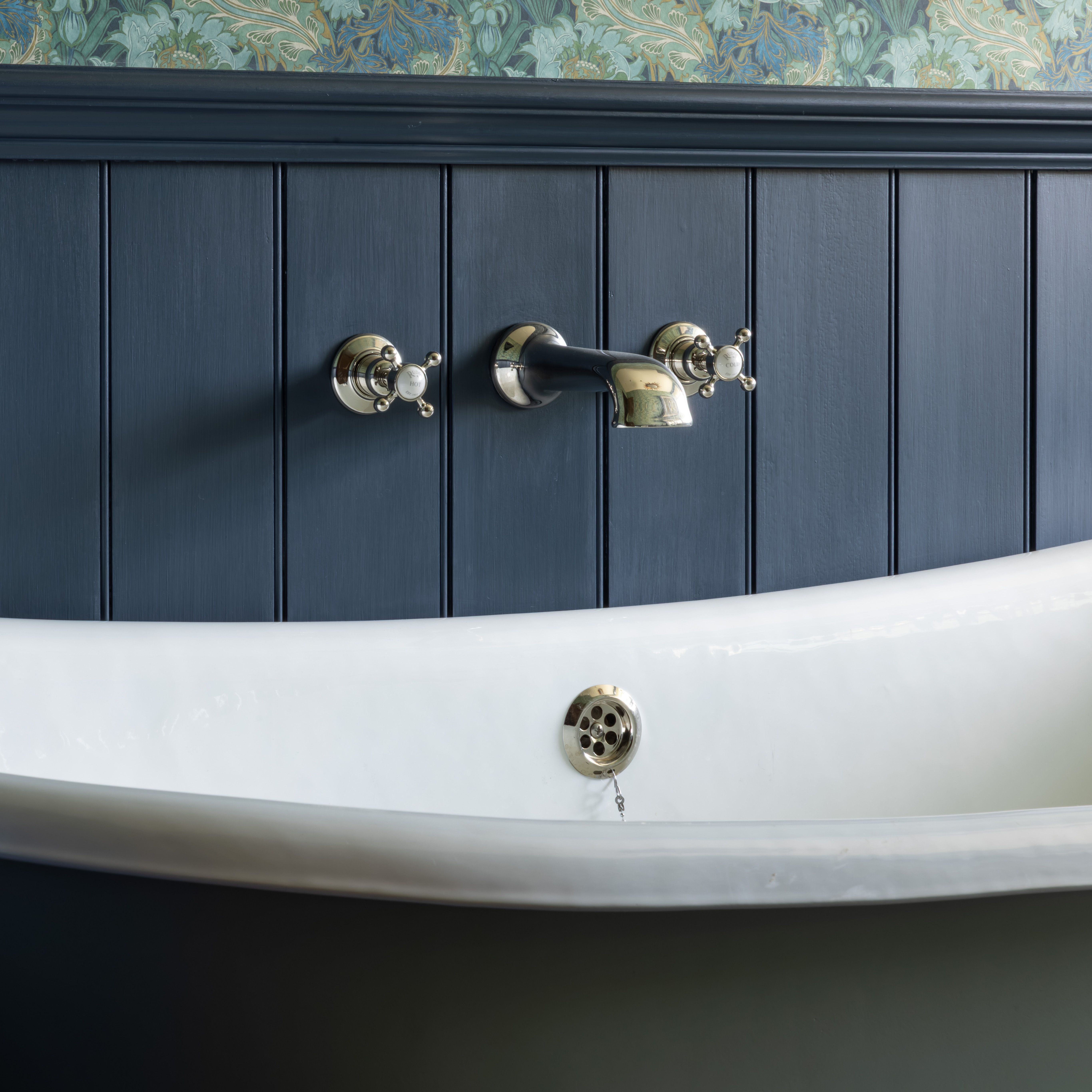 Close-up of a Victorian-style bathtub with vintage chrome fixtures against navy paneling and floral wallpaper.