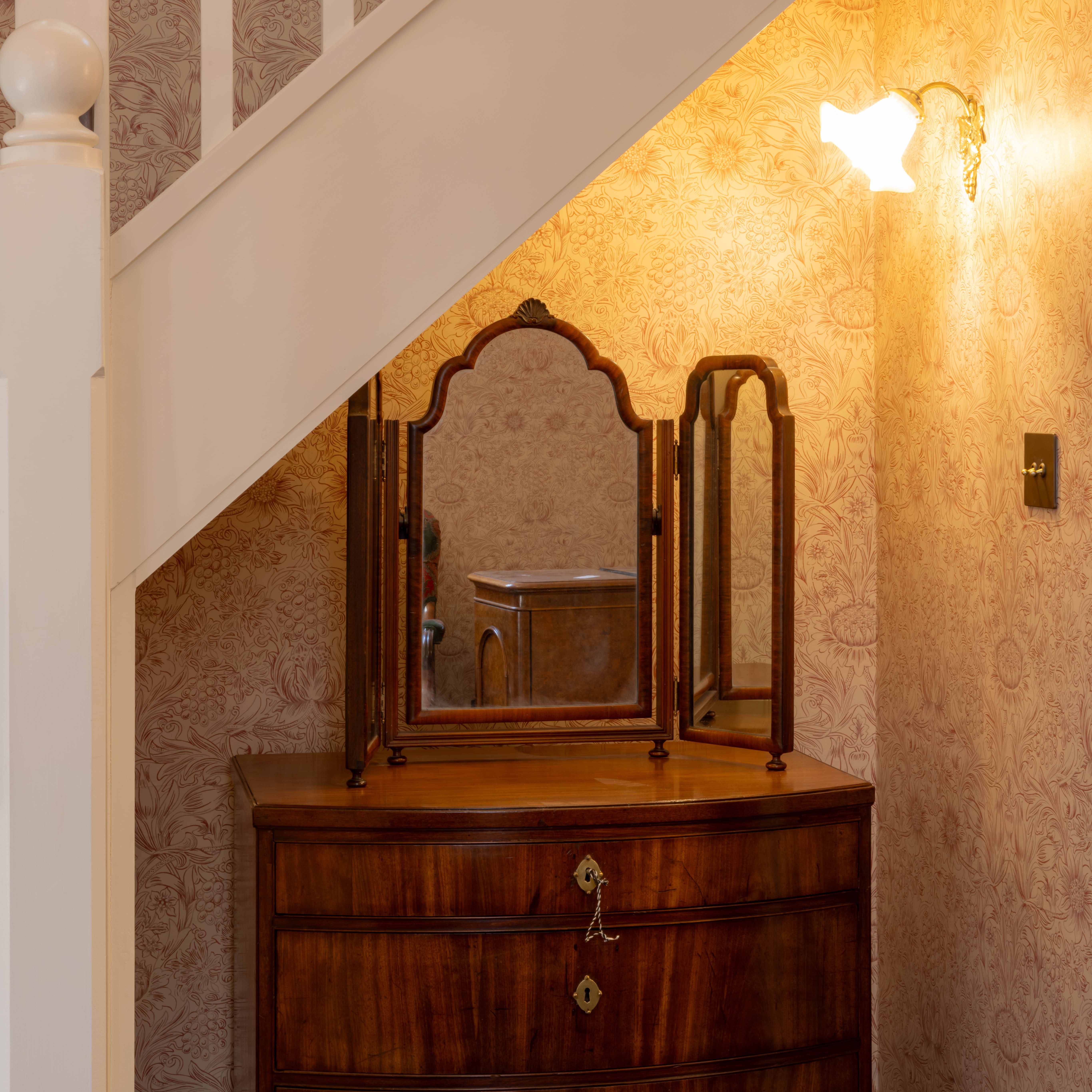 Victorian-style corner with floral wallpaper, a wooden chest of drawers with a three-paneled mirror under a staircase, and a lit wall sconce.