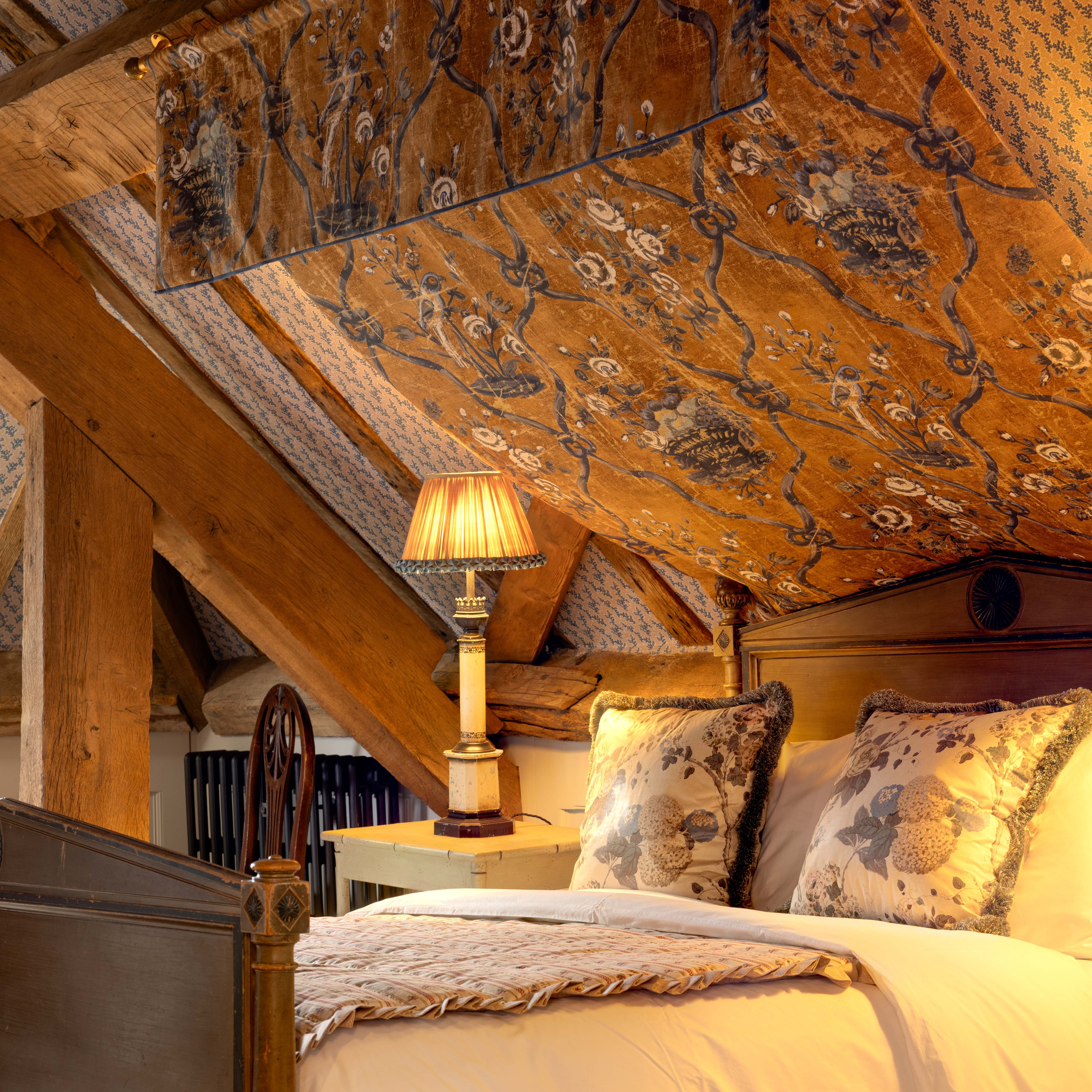 Cozy attic bedroom with a wooden Captains bed, floral pillows, and a vintage lamp under a sloped, patterned ceiling.