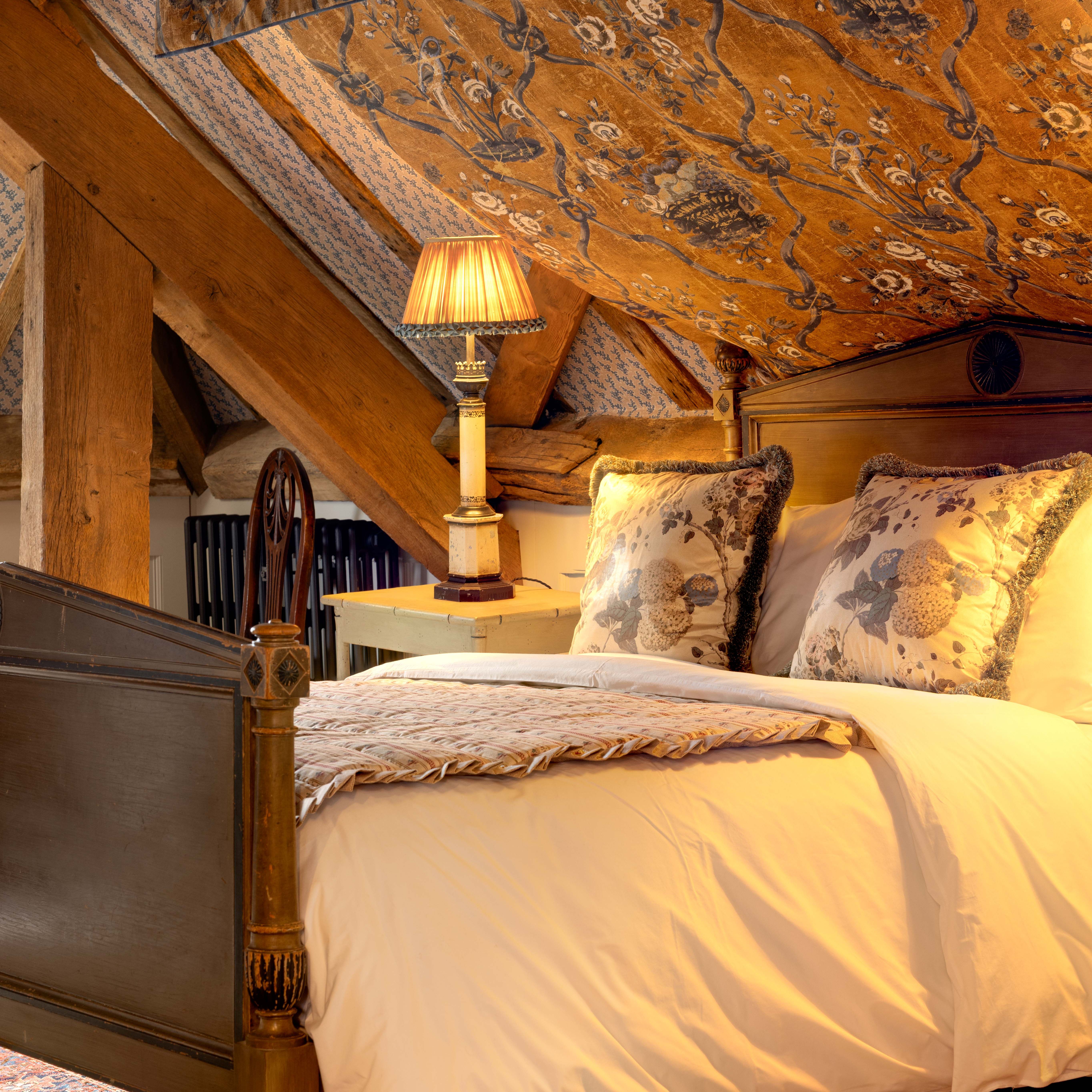 Cozy attic bedroom with a wooden Captains bed, floral pillows, and a vintage lamp under a sloped, patterned ceiling.