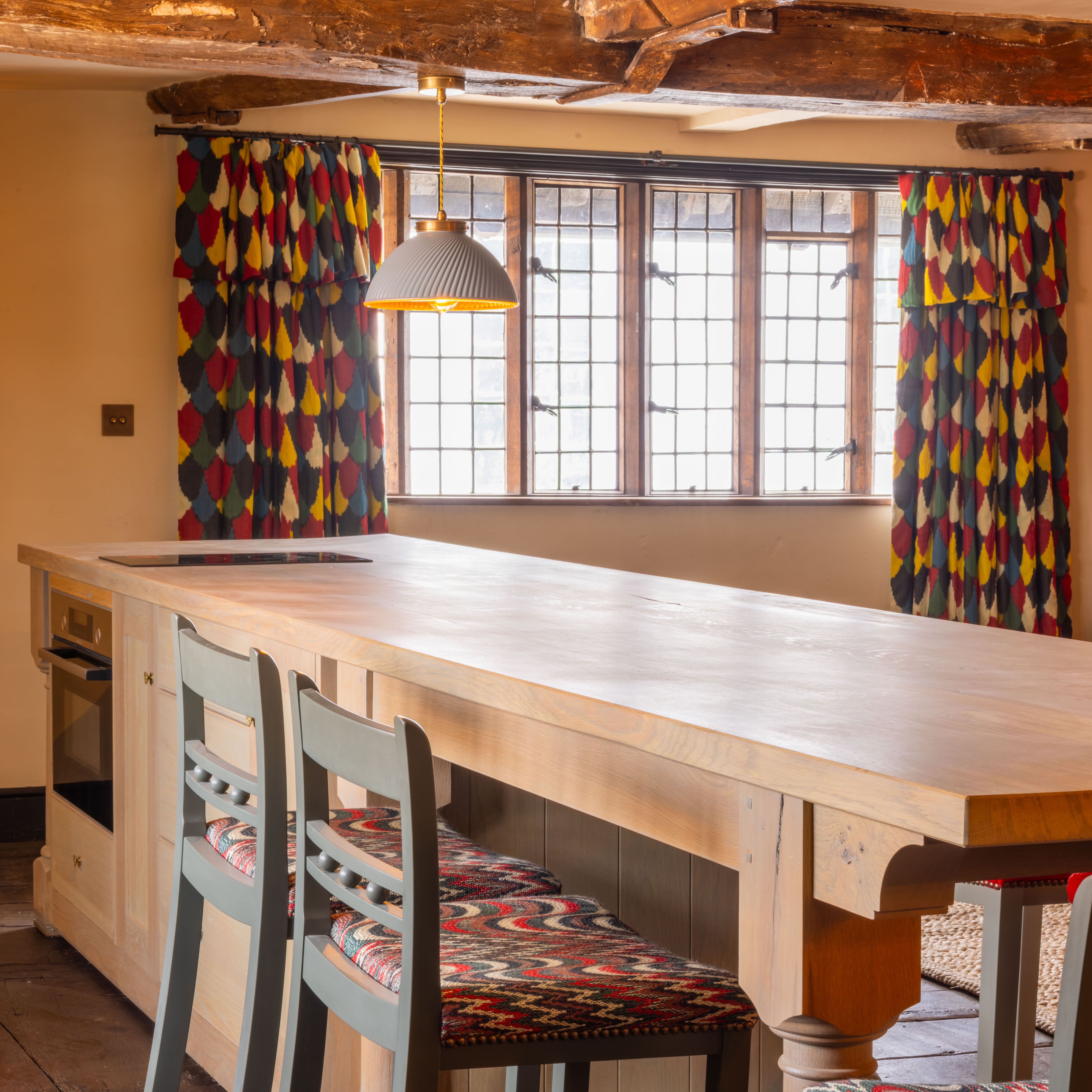 Large wooden banquet table in a rustic kitchen with colorful curtains and wooden beams