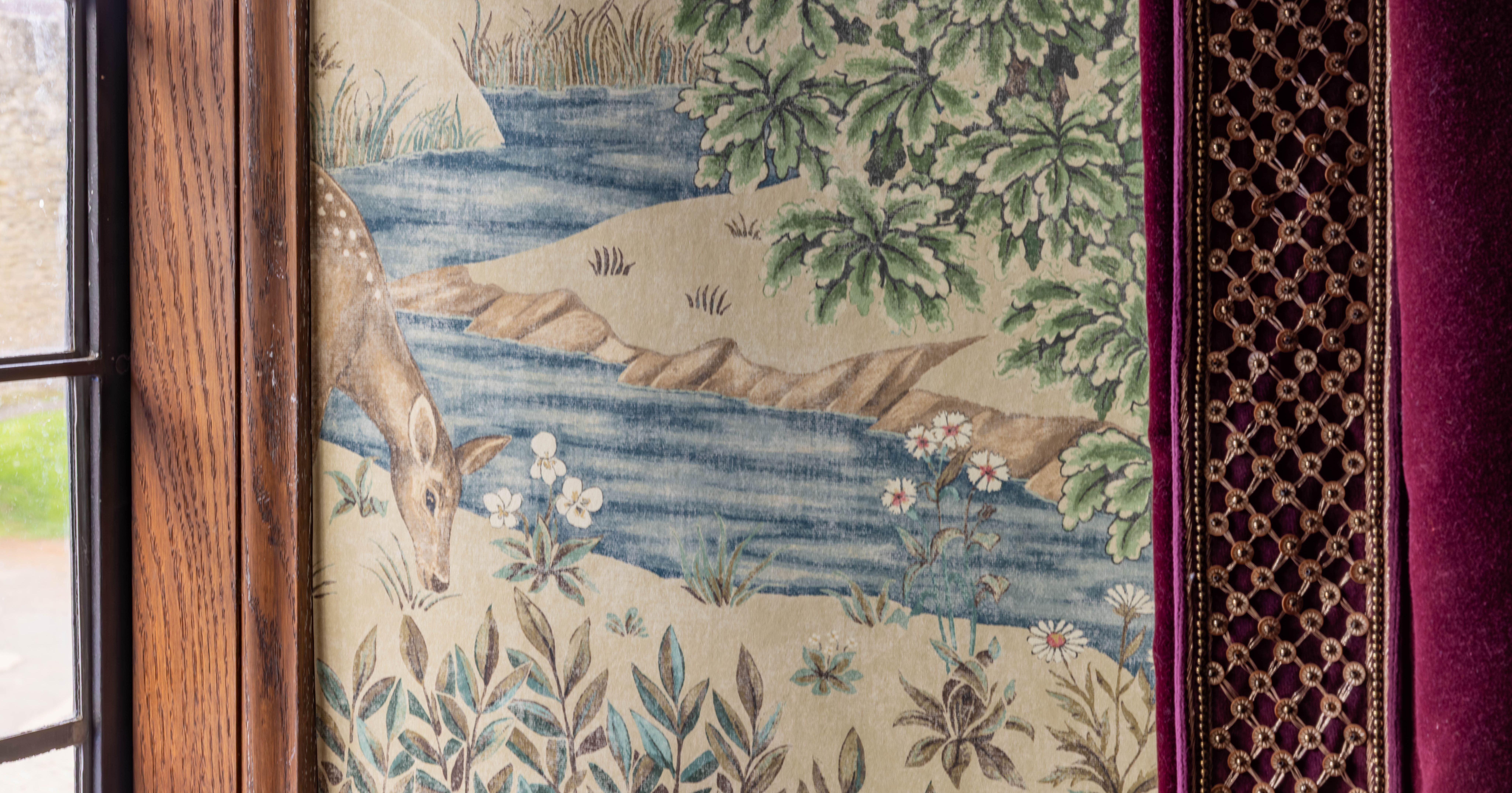 historic wallpaper featuring a deer by a stream with foliage and flowers next to a window and velvet curtain