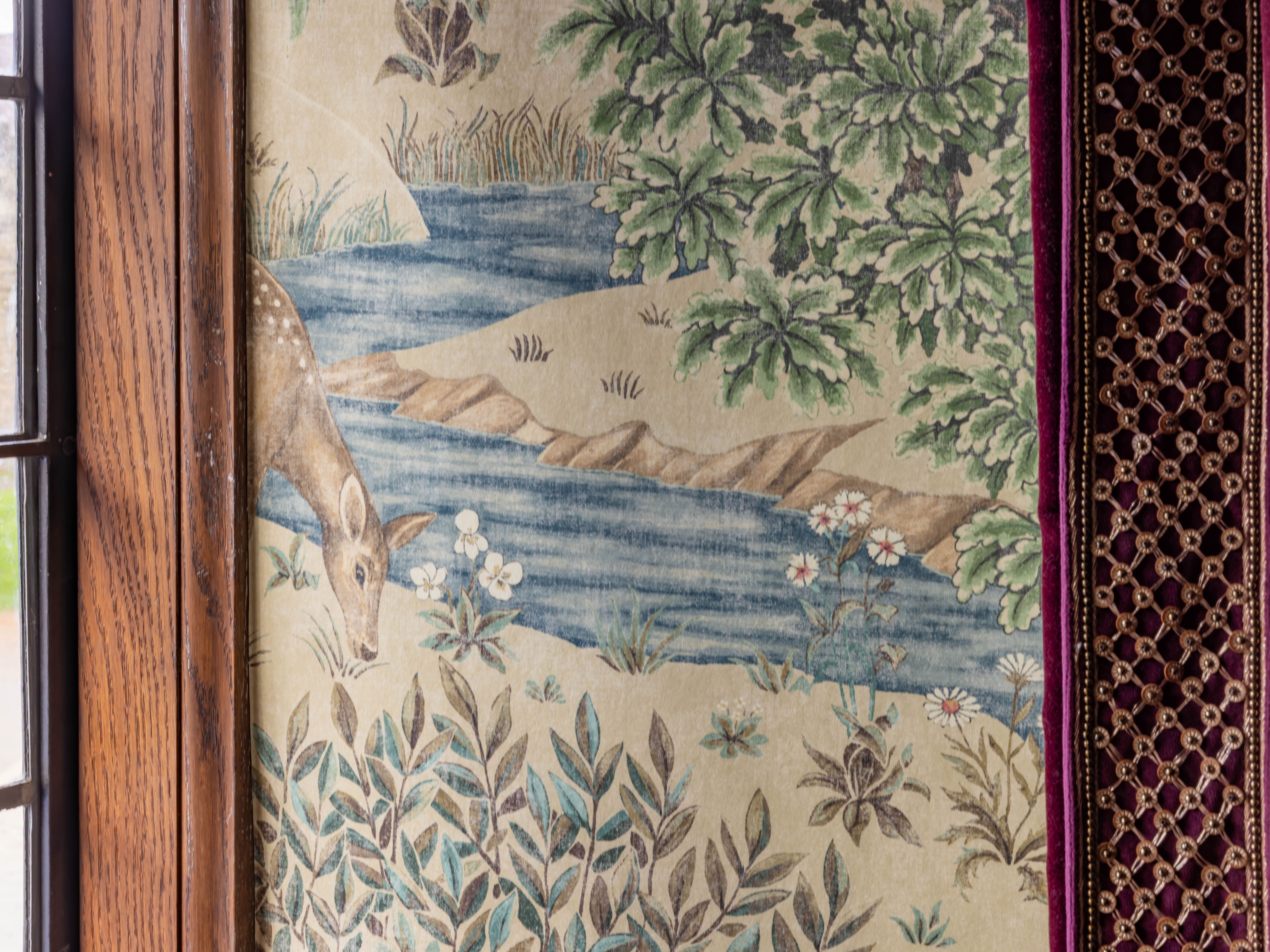 historic wallpaper featuring a deer by a stream with foliage and flowers next to a window and velvet curtain
