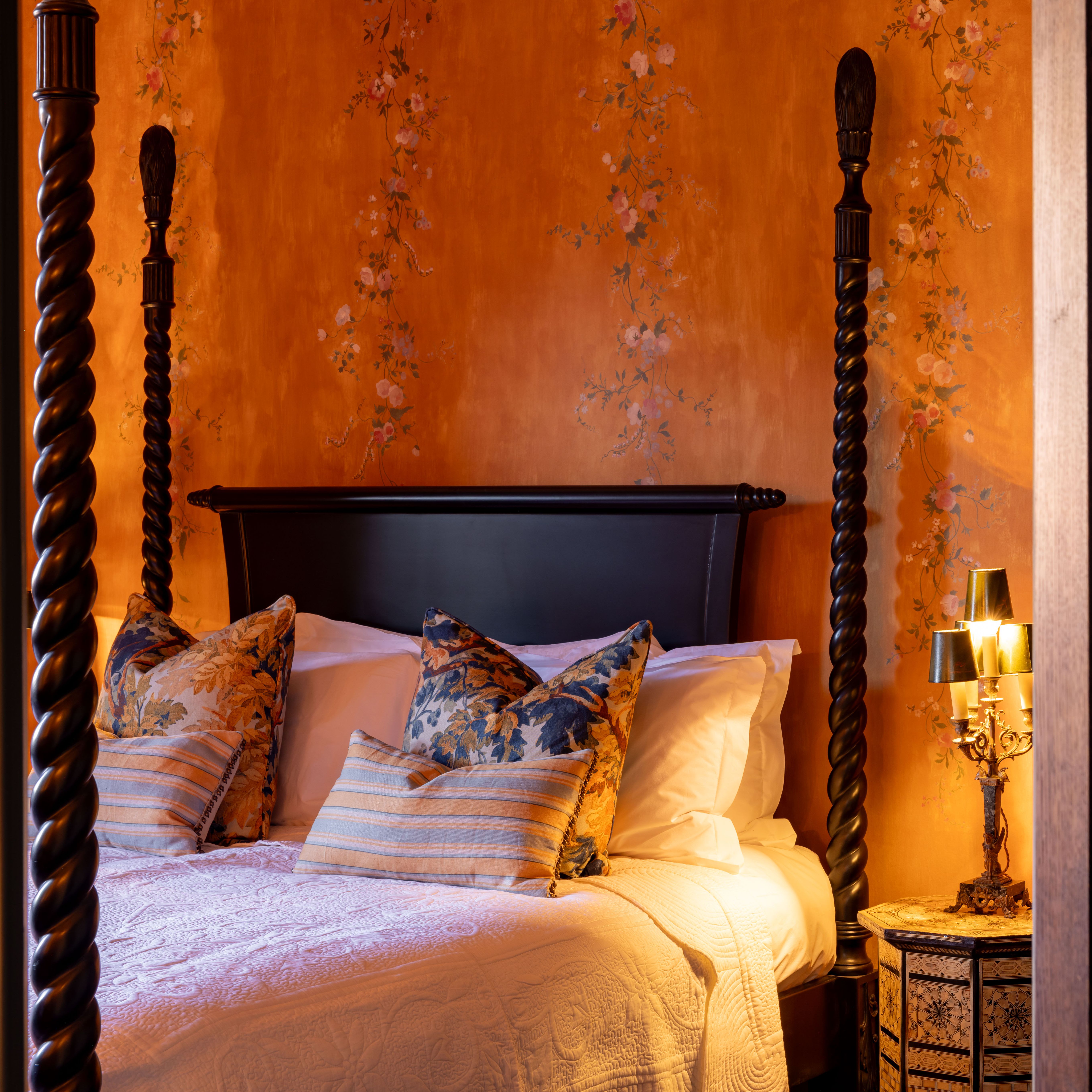 Elegant four-poster bed in a warmly lit bedroom with floral wallpaper and decorative pillows