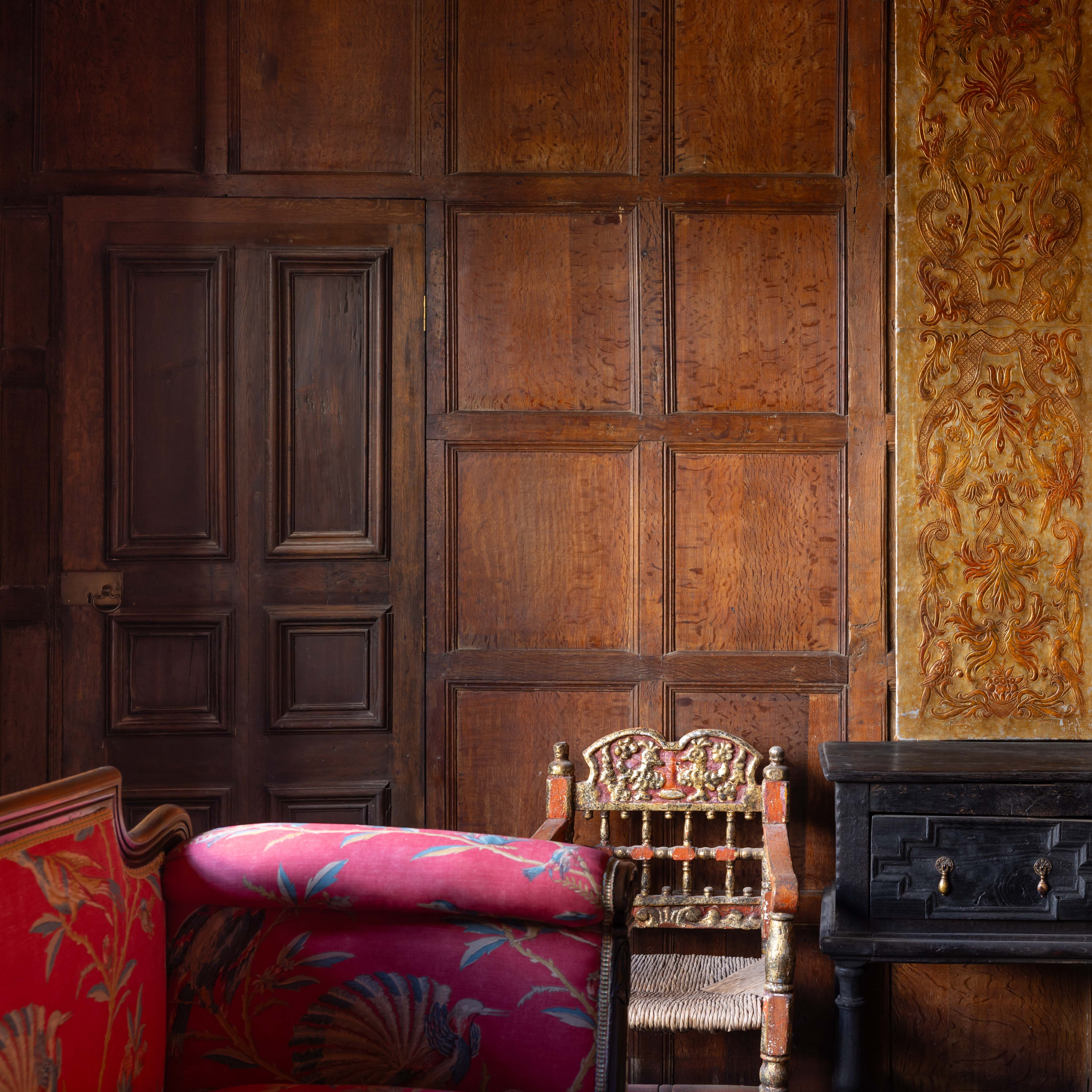 Historic lounge with ornate wooden paneling, antique chair, red upholstered sofa, and decorated cabinet.