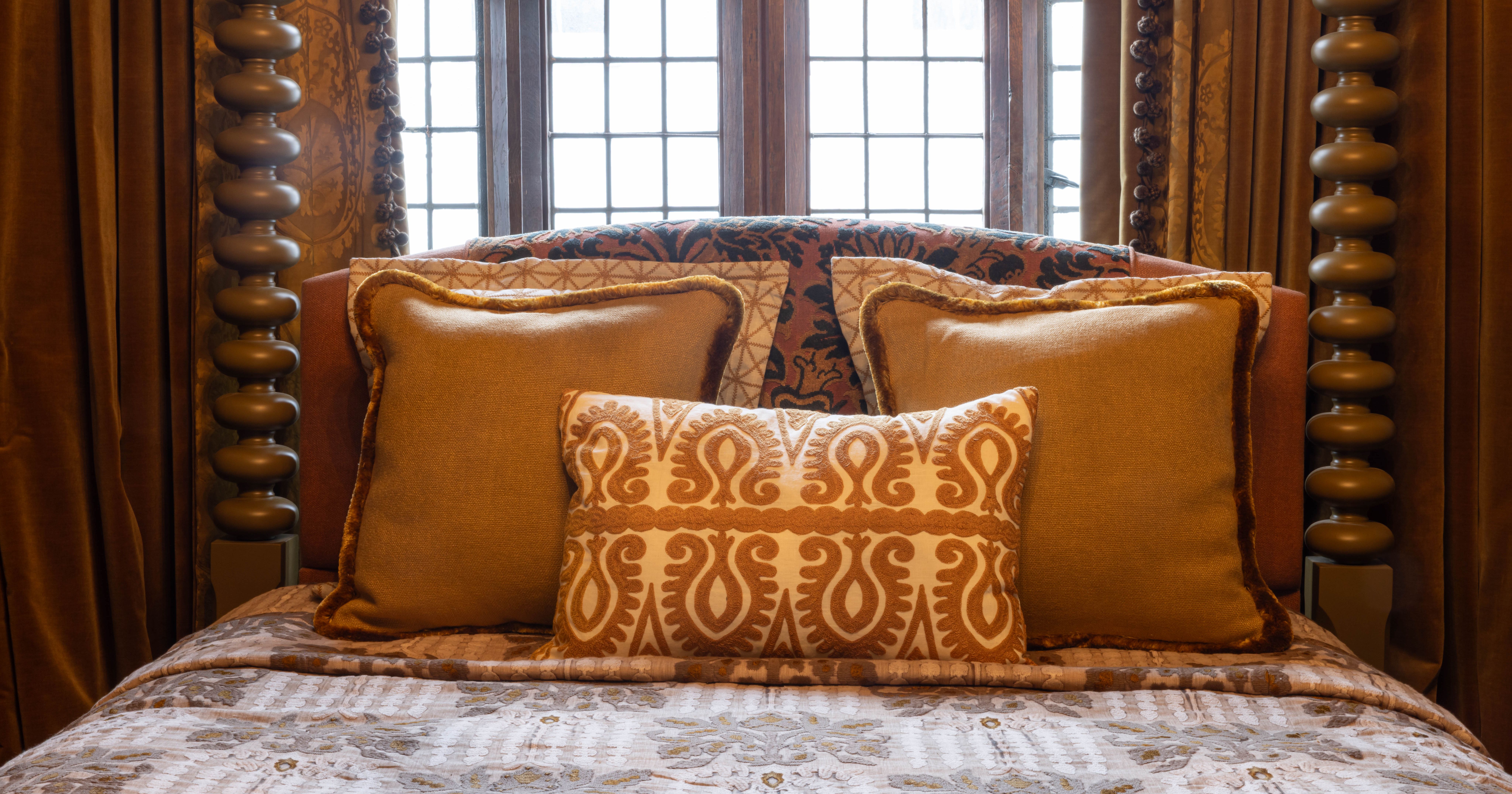 Close up of a luxurious bed with ornate pillows and an antique wooden headboard set against a leaded glass window.