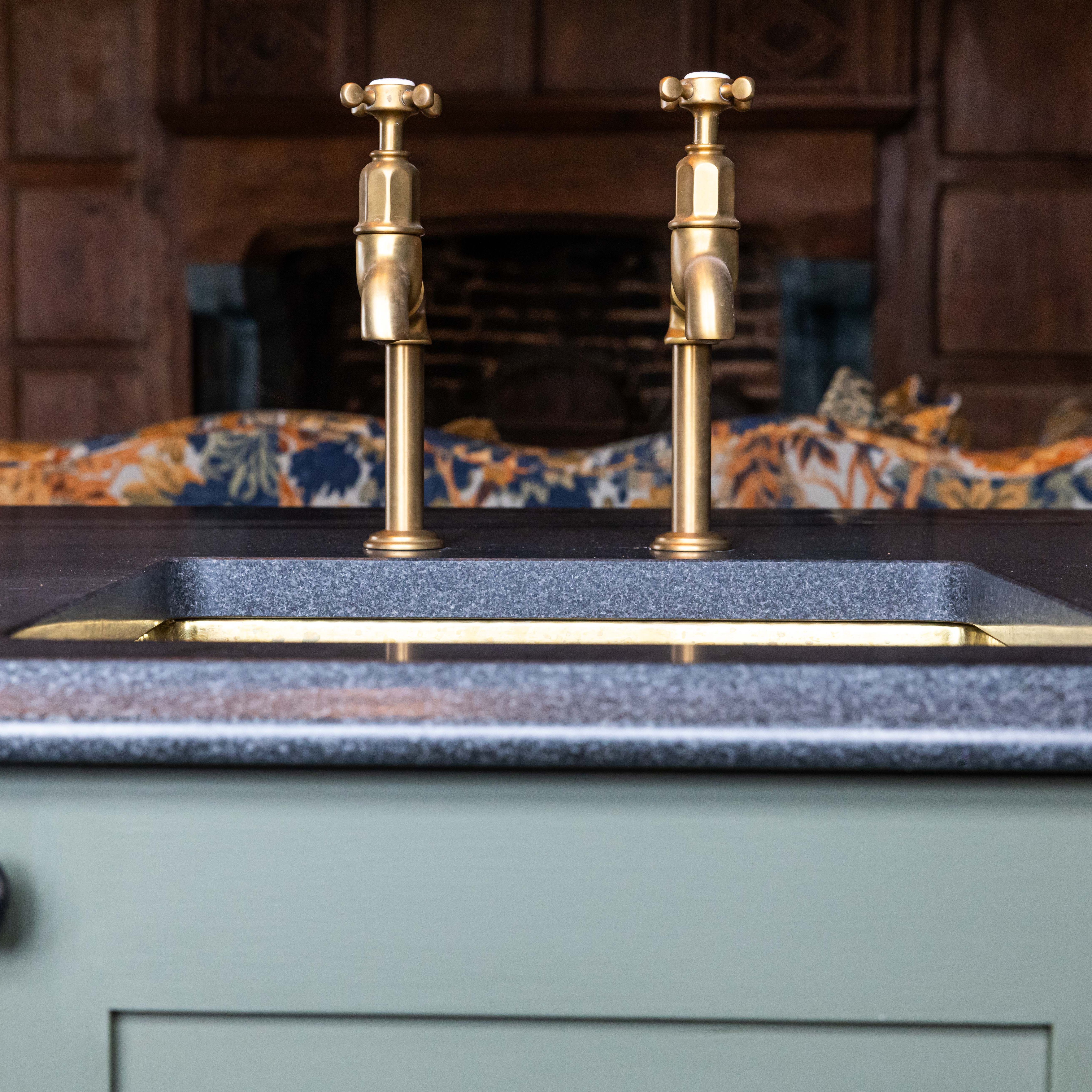 Close-up of a double brass kitchen tap over a dark countertop with a gold-edged sink, set against a vintage wood-paneled background.