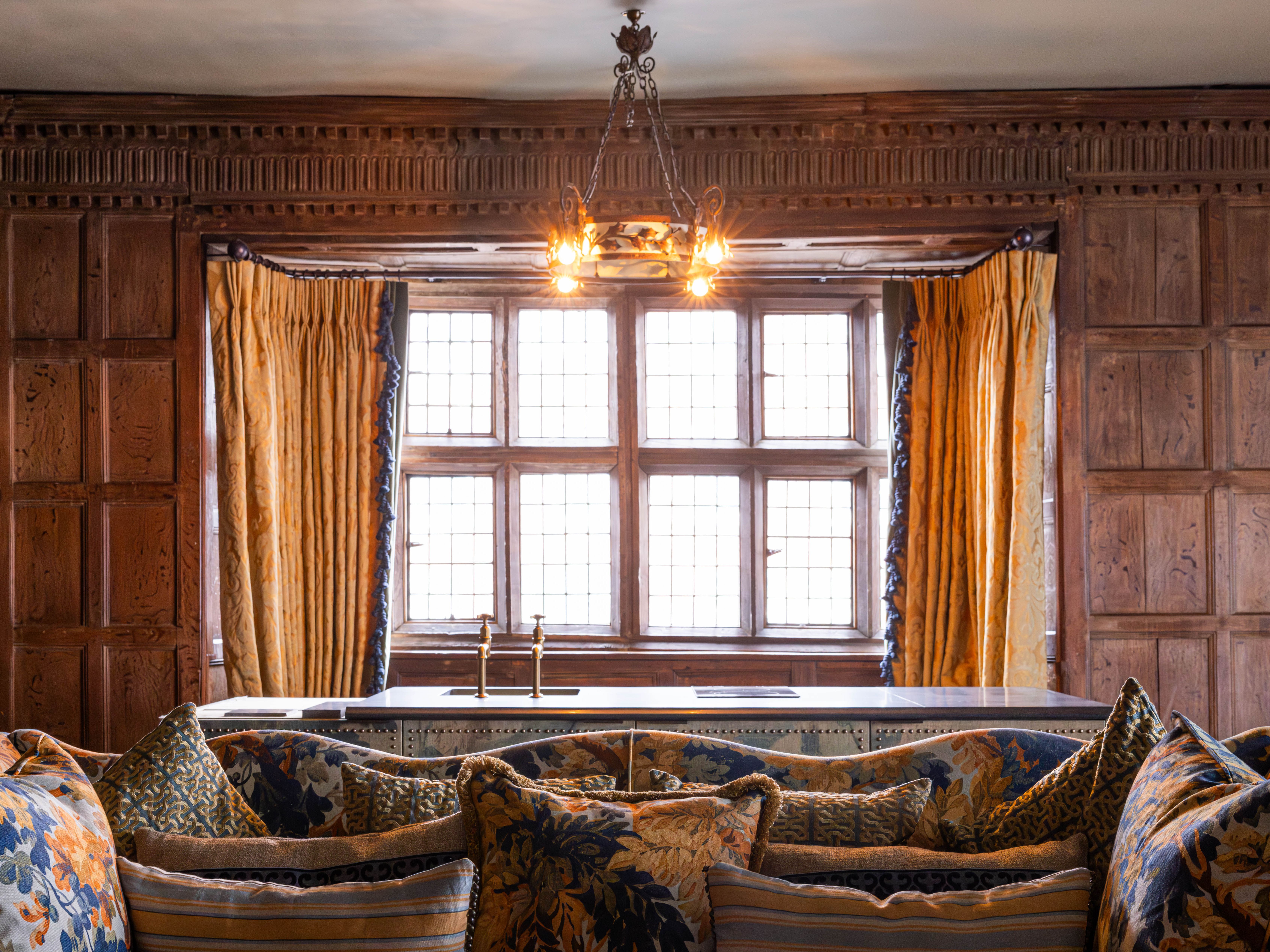 Elegant historic lounge with wood-paneled walls, large leaded glass windows, ornate gold curtains, a vintage chandelier, and a plush sofa with patterned cushions.