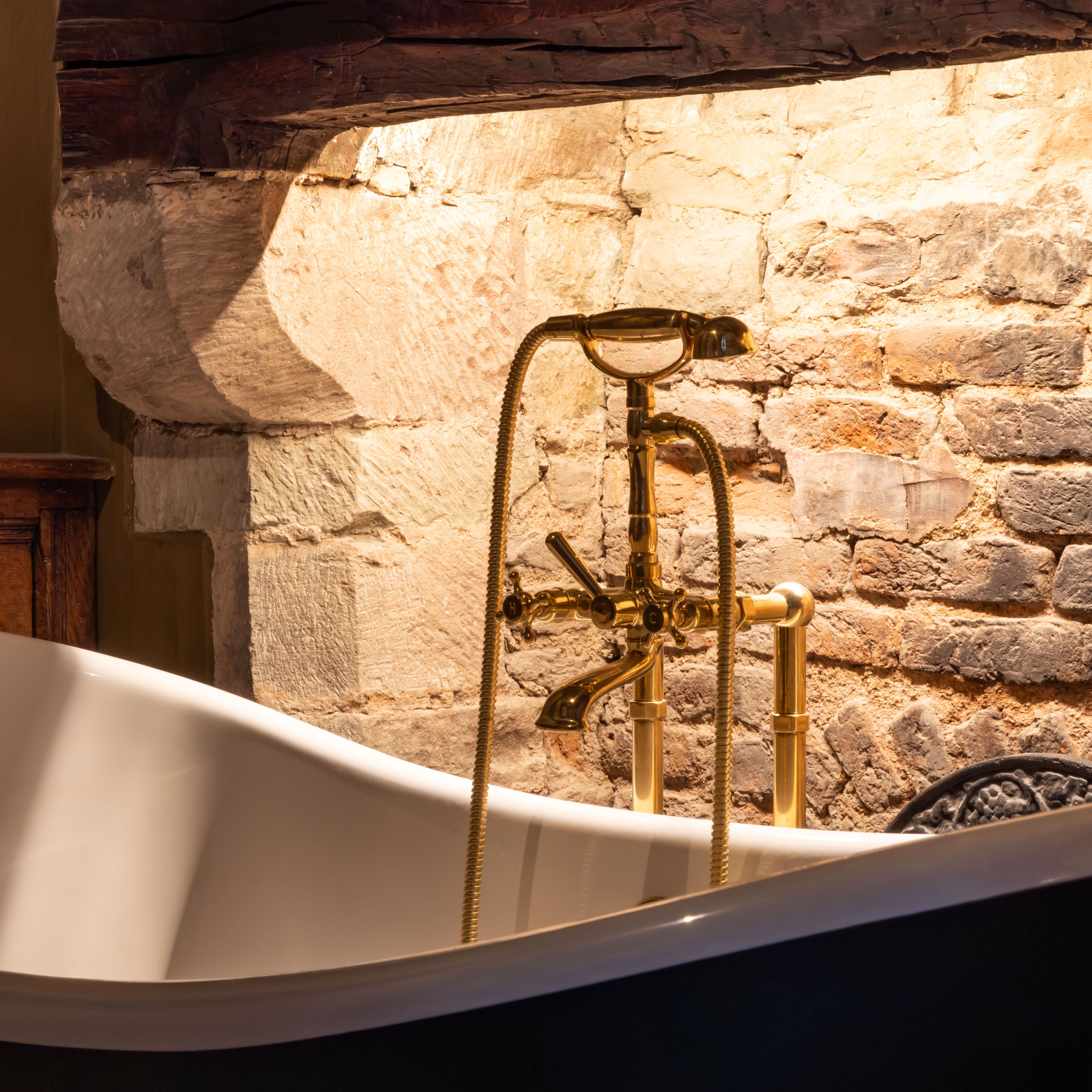 Vintage bathtub with gold faucet in front of exposed brick wall and wooden beam.