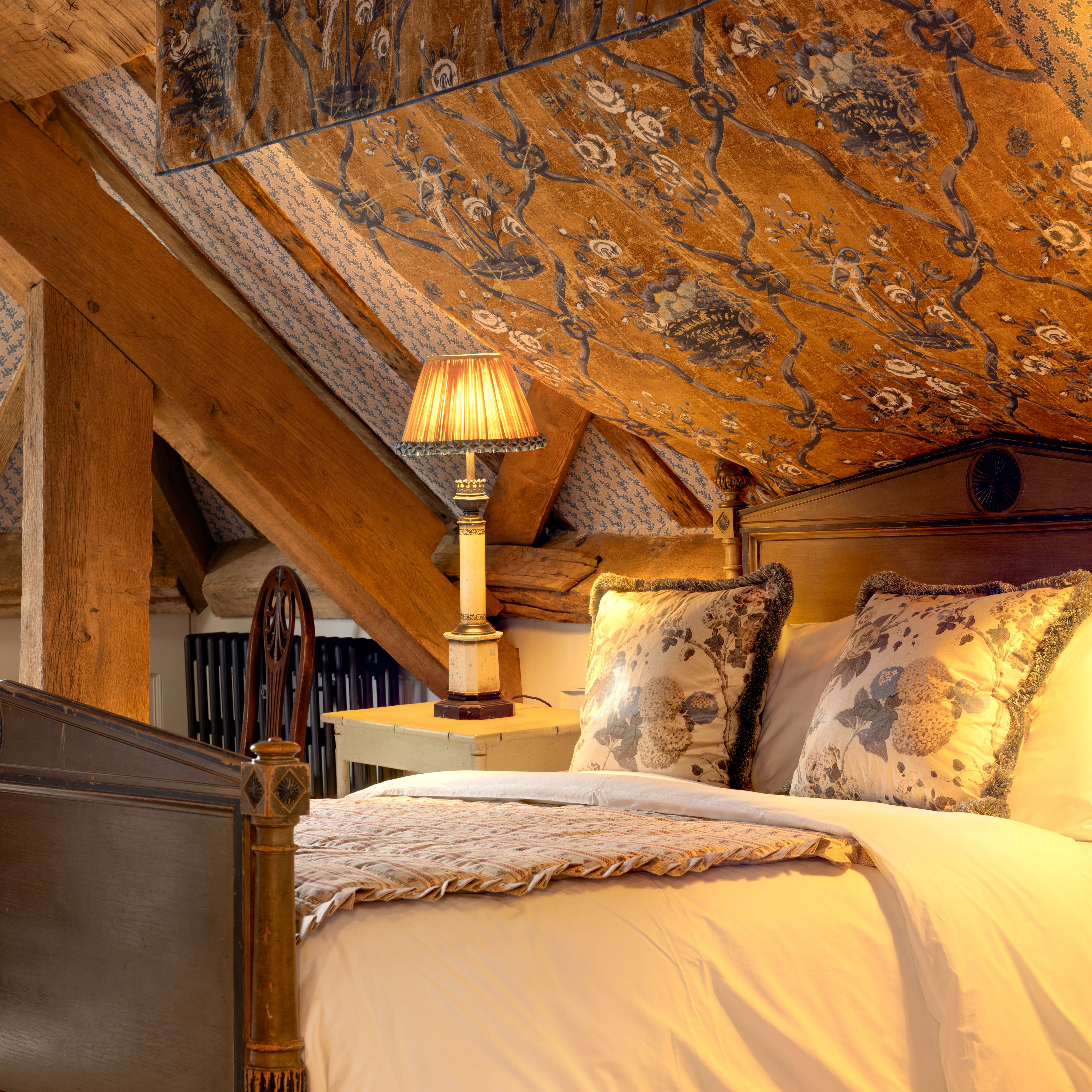 Cozy attic bedroom with a wooden captains bed, decorative pillows, patterned ceiling, and soft lighting from a bedside lamp.