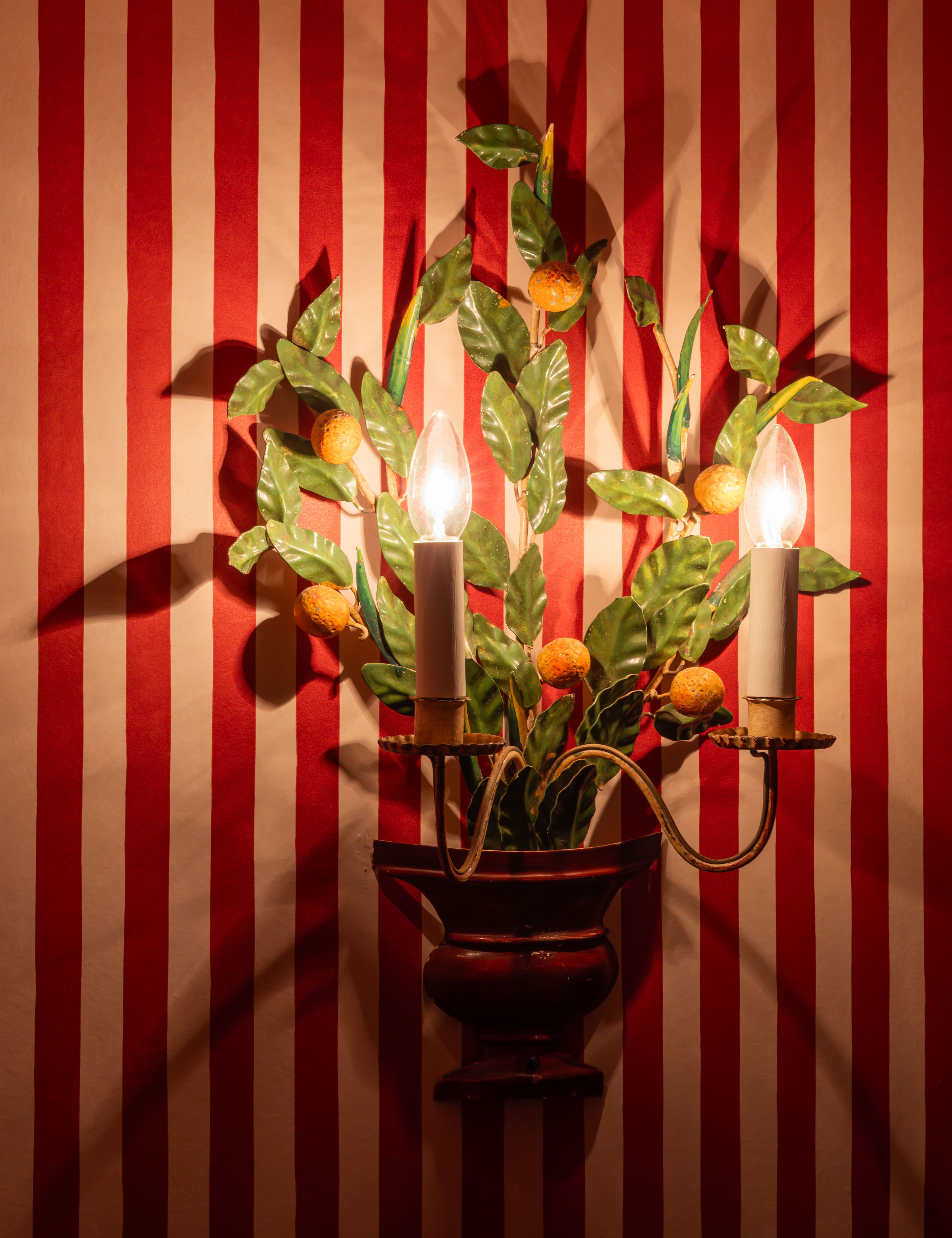 Decorative wall sconce with two lights shaped like candles and leaves, mounted on red and white striped wallpaper.