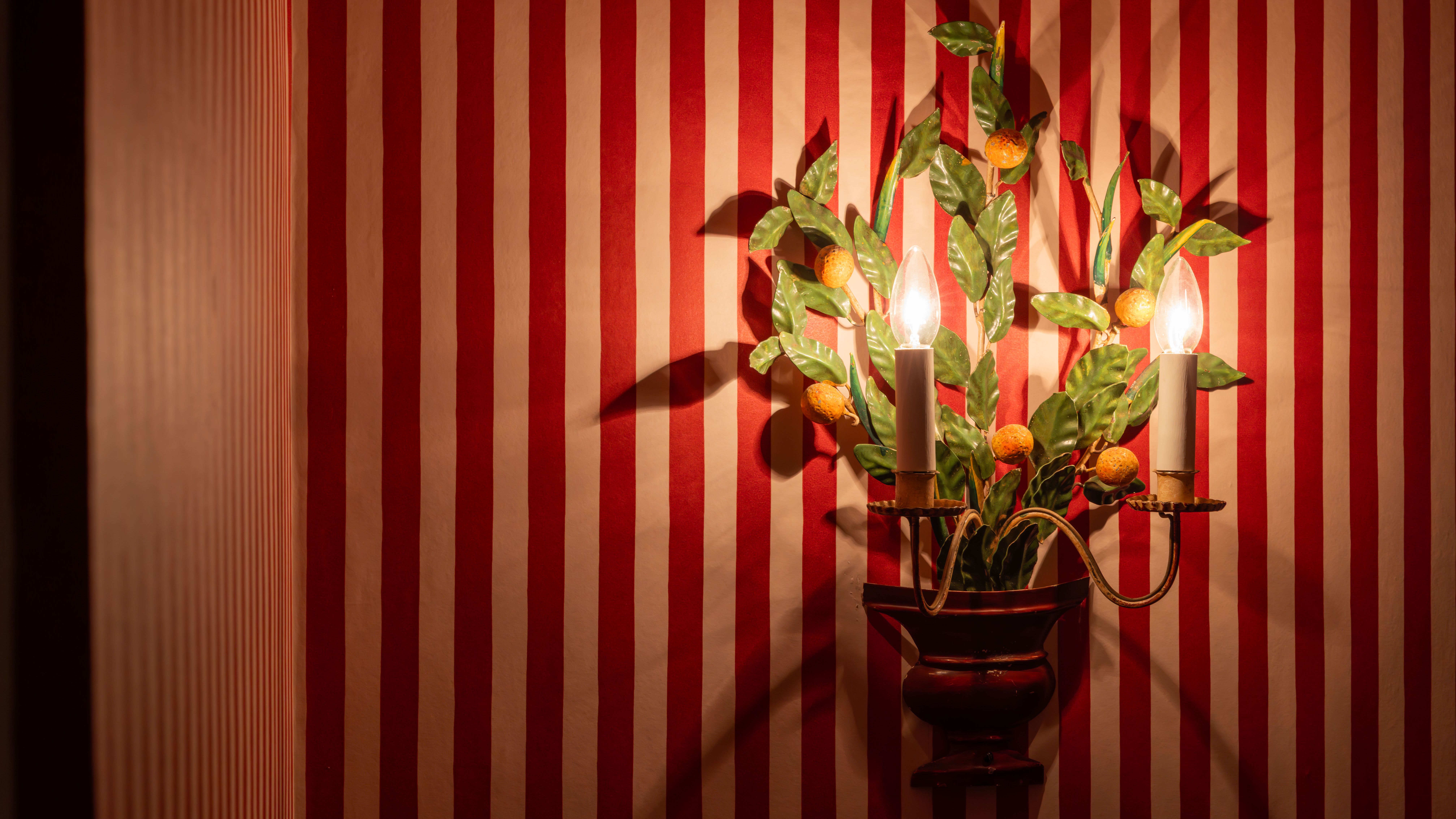 Decorative wall sconce with two lights shaped like candles and leaves, mounted on red and white striped wallpaper.