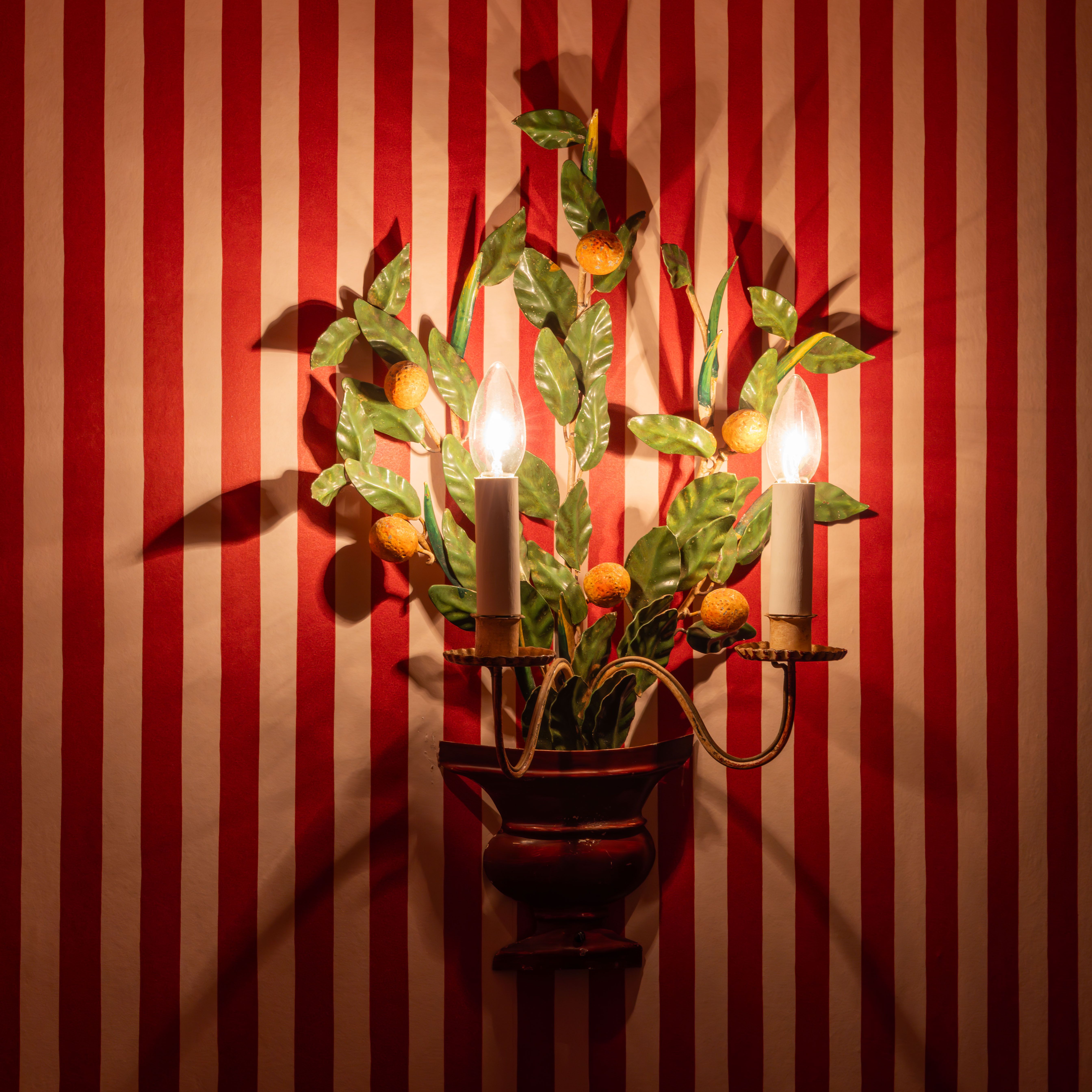 Decorative wall sconce with two lights shaped like candles and leaves, mounted on red and white striped wallpaper.
