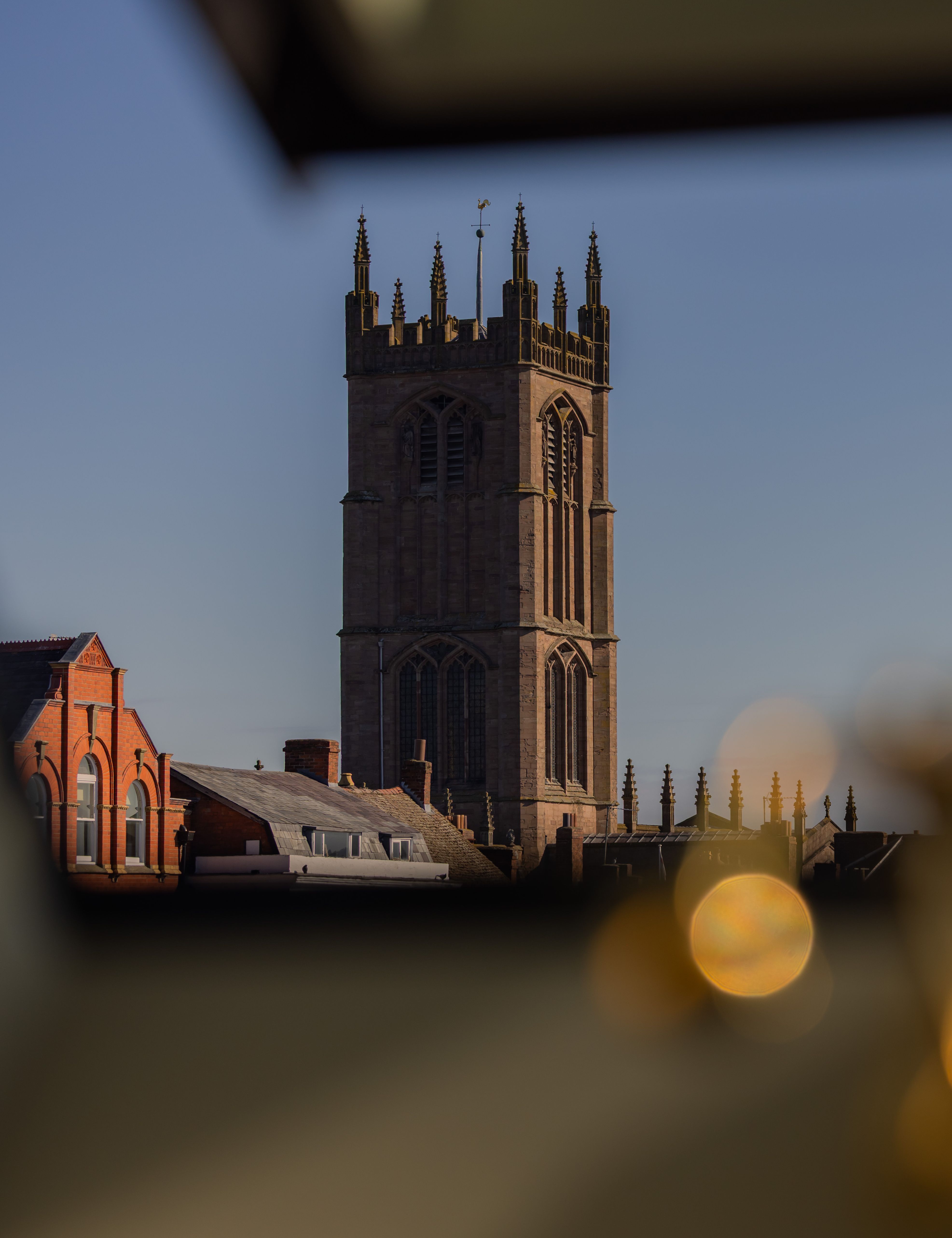 A view through a window showing a tall stone church tower, surrounding rooftops, and distant hills under a clear sky.