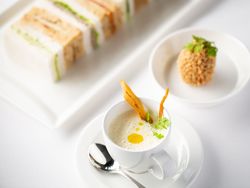 Elegant afternoon tea set with a cup of soup, finger sandwiches, and a breaded appetizer on white dishes.