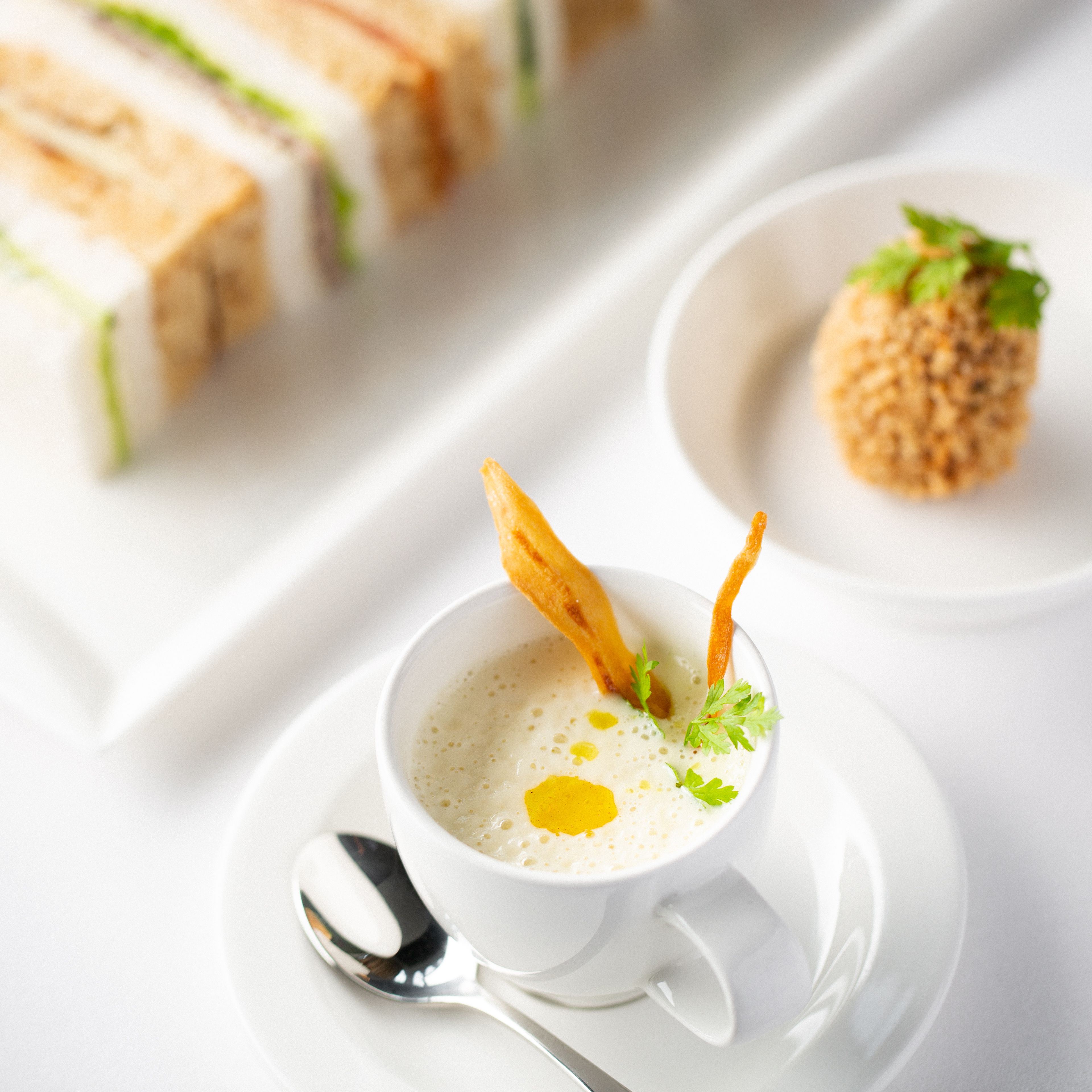 Elegant afternoon tea set with a cup of soup, finger sandwiches, and a breaded appetizer on white dishes.