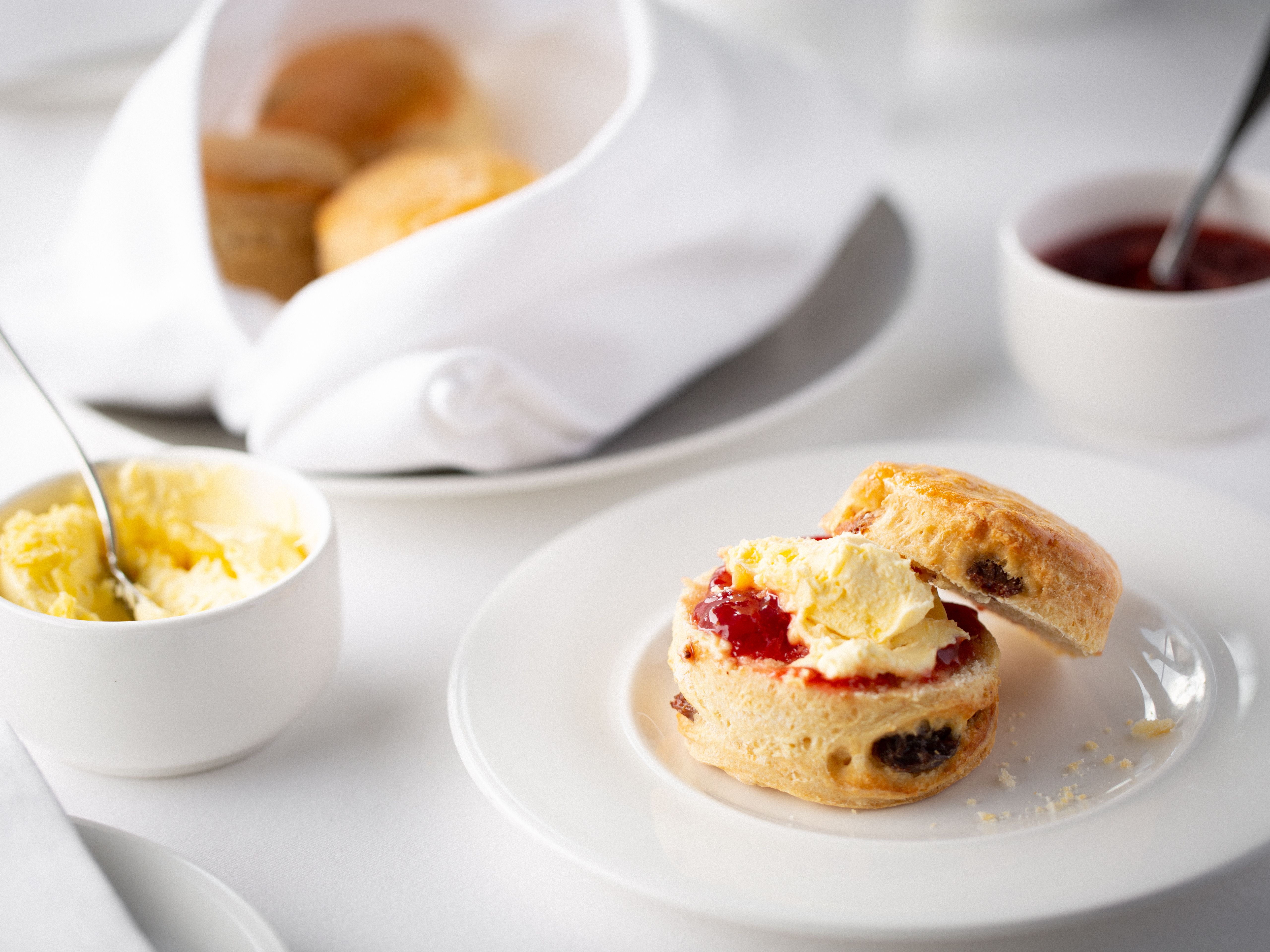Scone with clotted cream and strawberry jam on a white plate, butter and jam in small bowls, with more scones in a napkin-lined basket.