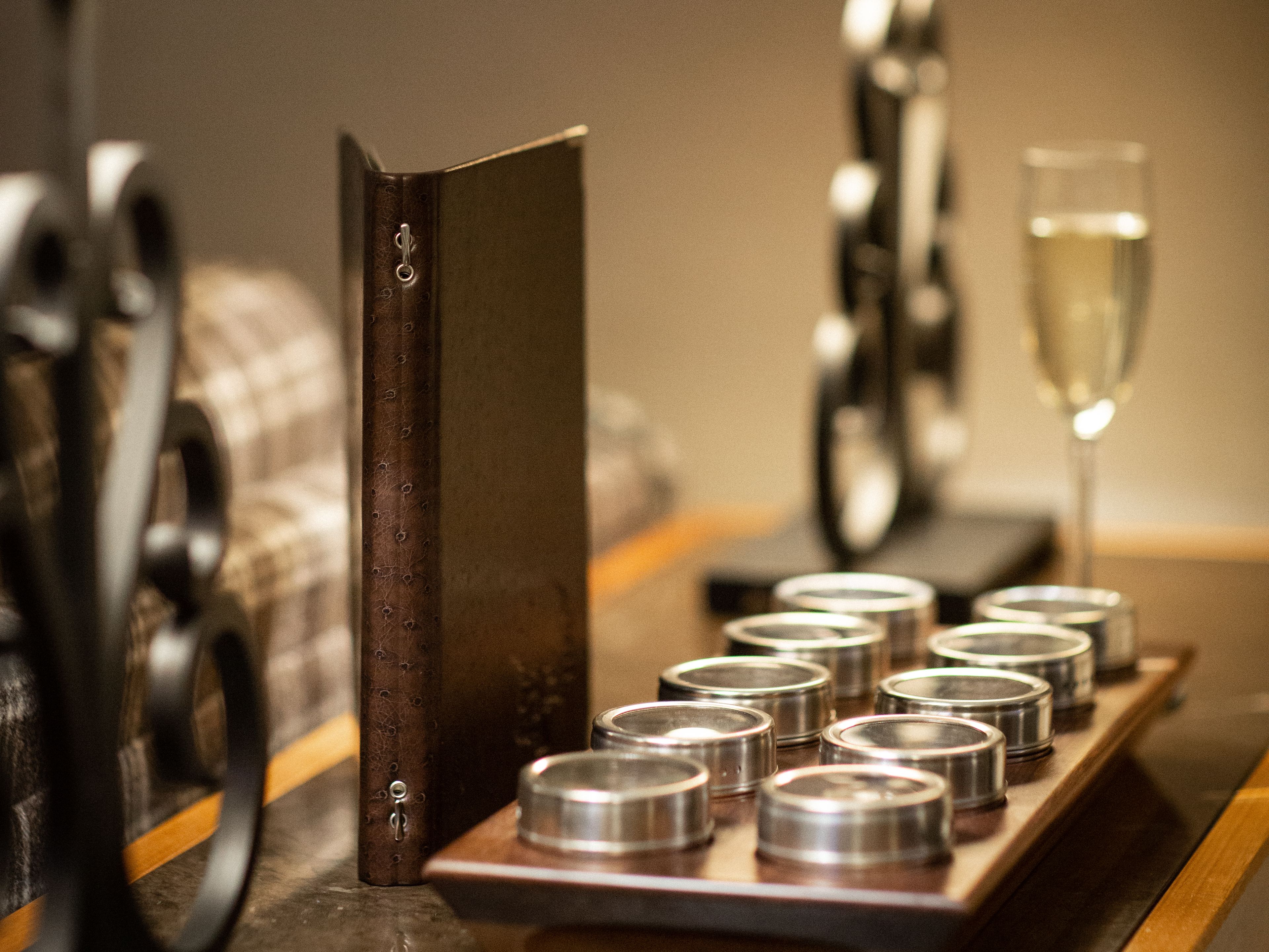 A cozy table setting with two lamps, a closed menu or book, a tray with small metal containers, and a glass of champagne.