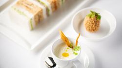 A gourmet appetizer set featuring a cup of creamy soup with garnish, a coated bite-sized treat in a small bowl, and assorted tea sandwiches on a white plate.