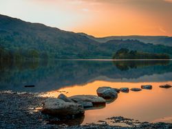 Serene lake with rocks in the foreground and hills in the background at sunset