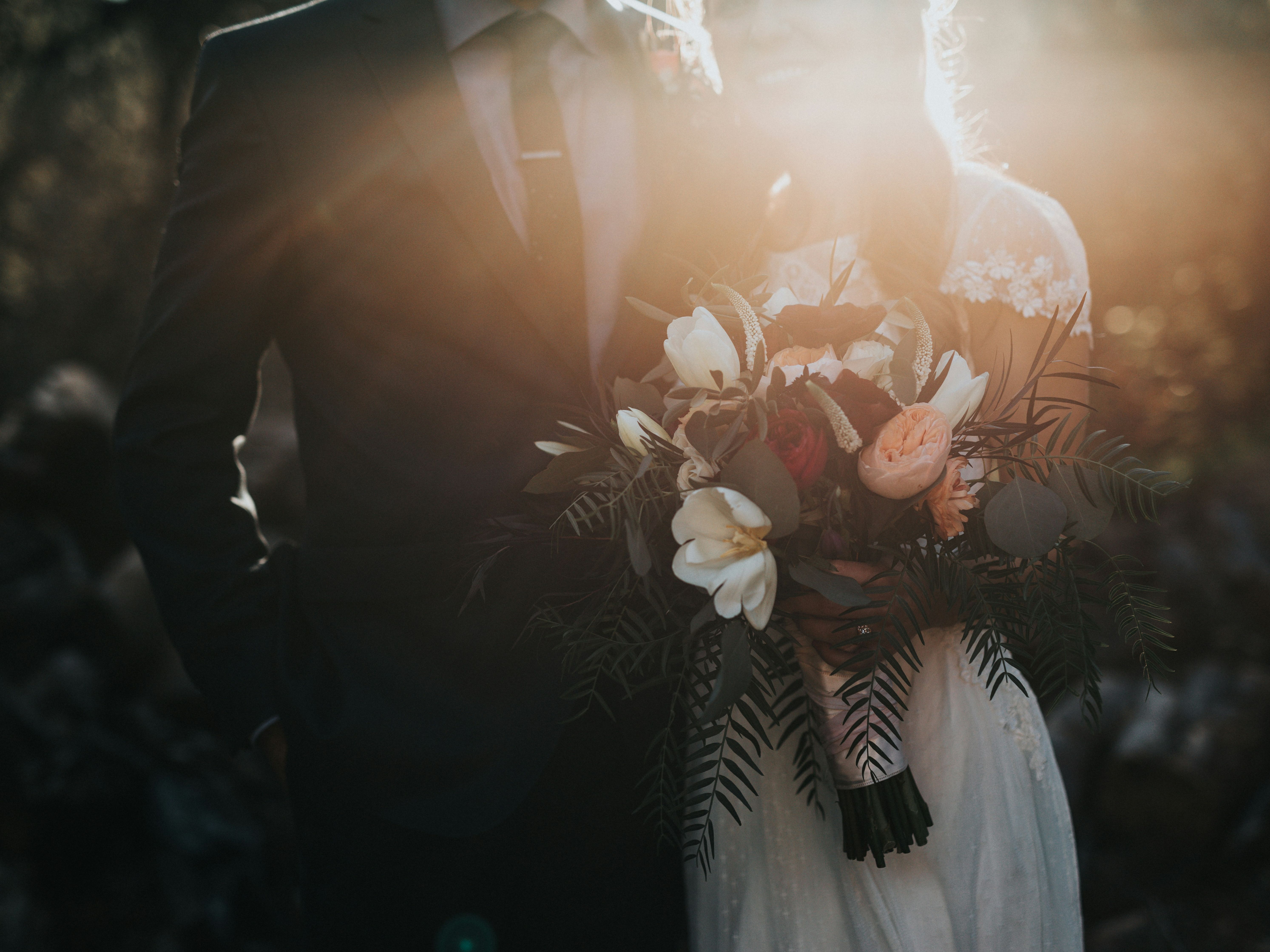 A bride and groom standing close together outdoors in soft sunlight, the bride holding a colorful bouquet of flowers.