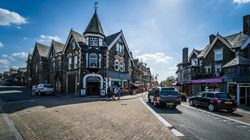 Street view of Windermere village with stone buildings, shops, and parked cars on a sunny day.